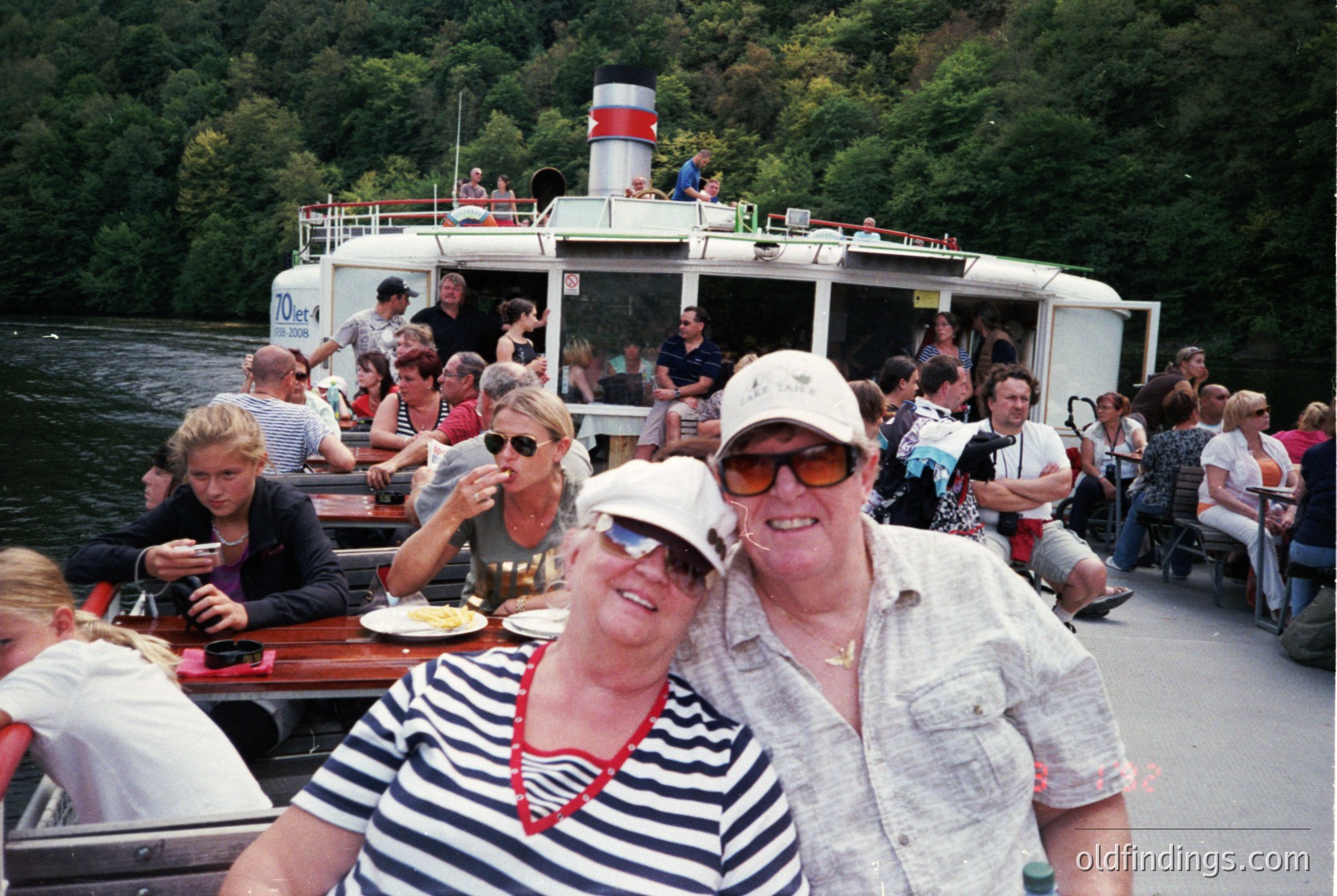 Classic 1970s-era river cruise boat packed with passengers enjoying a leisurely outing. Two smiling adults in retro sunglasses and striped shirts pose in the foreground, while others relax, eat, and socialize on deck. Lush green forest backdrop suggests a scenic lakeside or river setting.