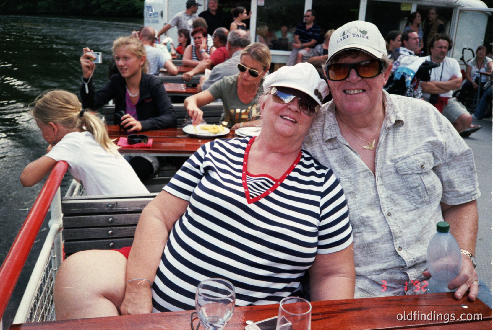 Two adults pose joyfully on a covered canal boat, likely a 1990s Dutch *taxi boot* (boat taxi). The man wears a light cap and sunglasses; the woman sports a striped shirt and red choker. Glasses, a water bottle, and casual summer attire suggest a leisurely, social outing. Crowded deck indicates popular tourist activity.