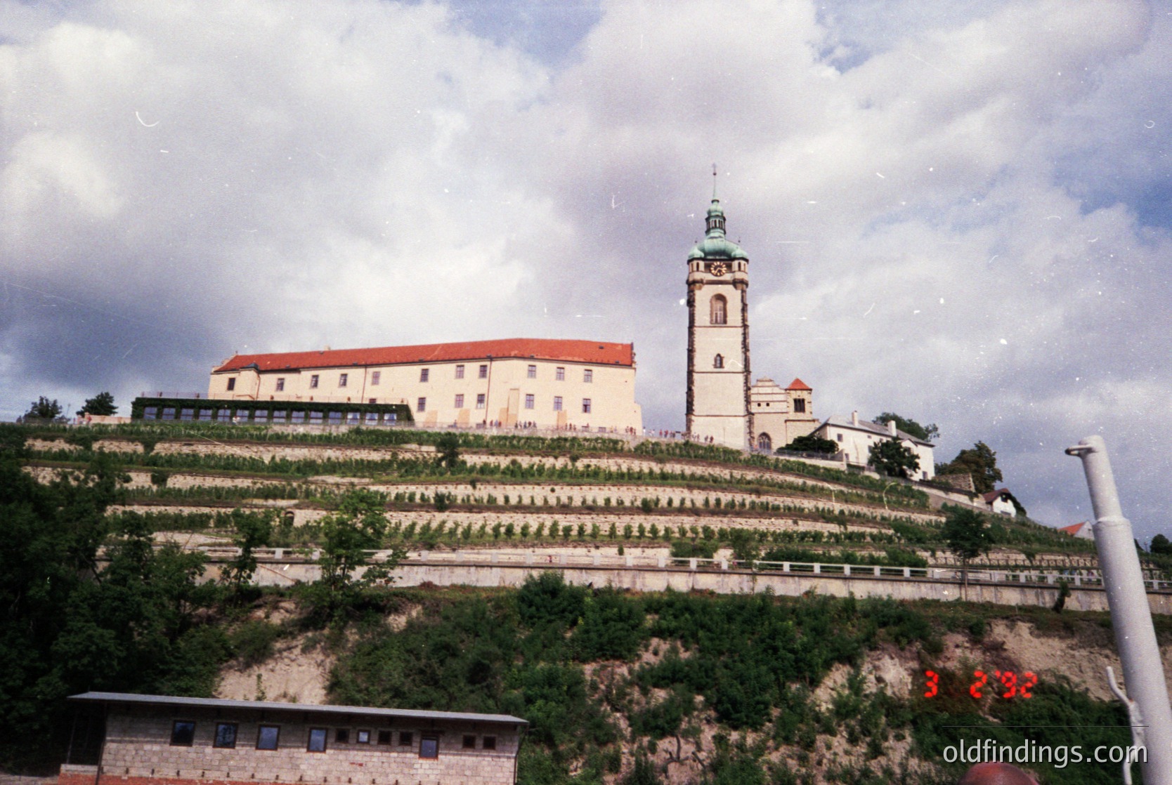 Historic hilltop monastery with red-tiled roof and central bell tower, surrounded by terraced greenery. Likely Eastern European architecture, possibly Bulgaria’s Rila Monastery. Mid-20th century vintage, showcasing religious heritage and landscape design.