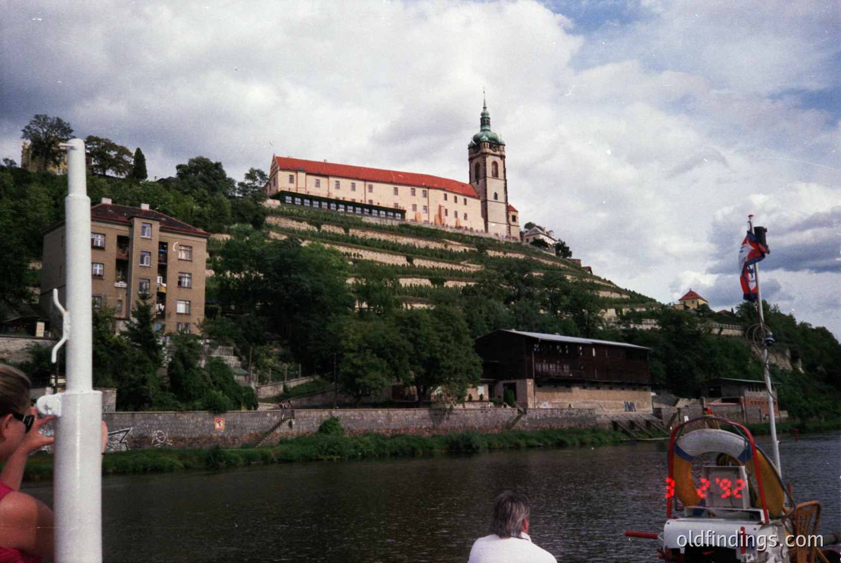 Historic castle perched on a hillside overlooking a river, featuring red-tiled roofs and a prominent tower. Mid-20th century urban architecture visible in foreground. Likely