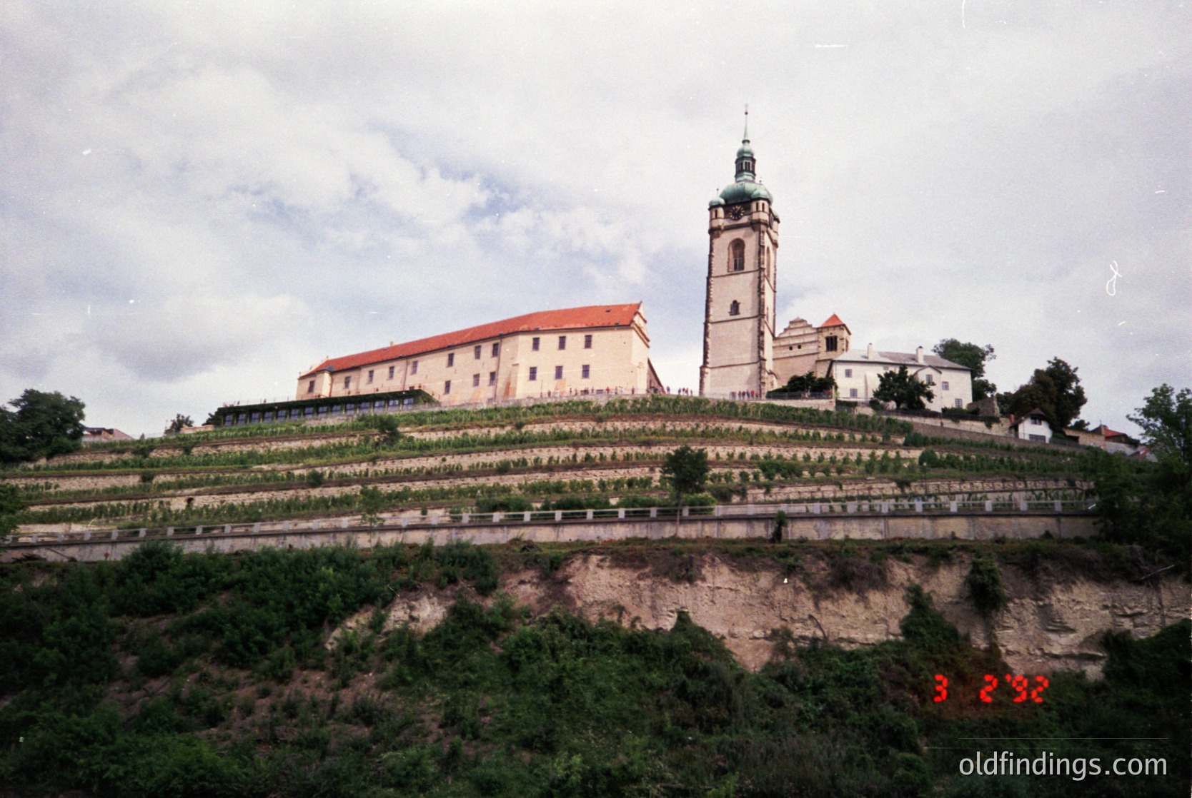 Historic monastery complex perched on terraced hillside, featuring a prominent bell tower with green dome and red-tiled roofs. Architectural style suggests Eastern European influence, likely Bulgaria. Lush greenery and stone pathways enhance the serene setting. Potential 1980s-1990s vintage based on film grain.