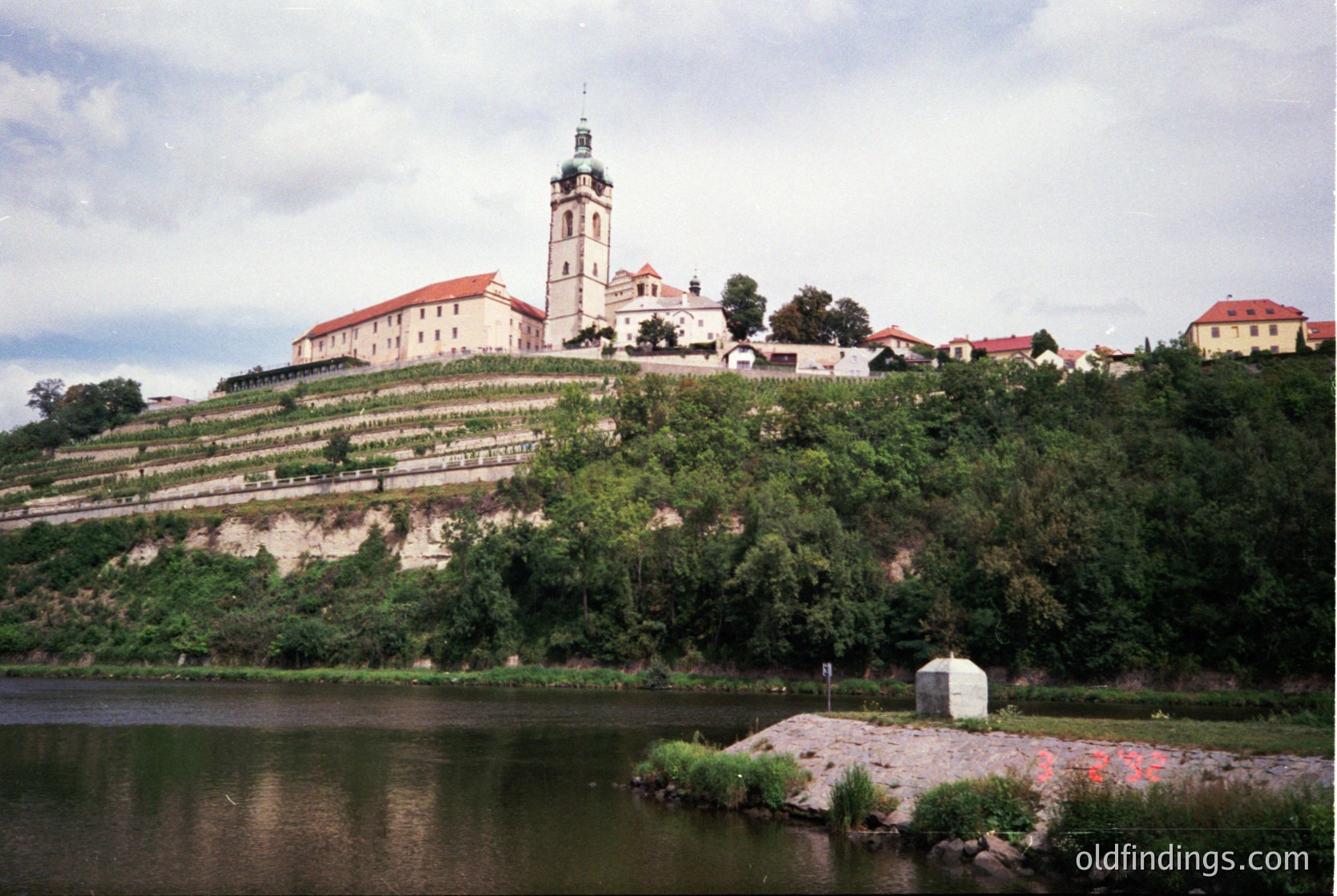 Historic hilltop fortress with Baroque-style church tower and vineyard terraces overlooking a river. Architectural details include red-tiled roofs and stone walls. Likely Central/Eastern European setting, possibly or . architecture with preserved elements.