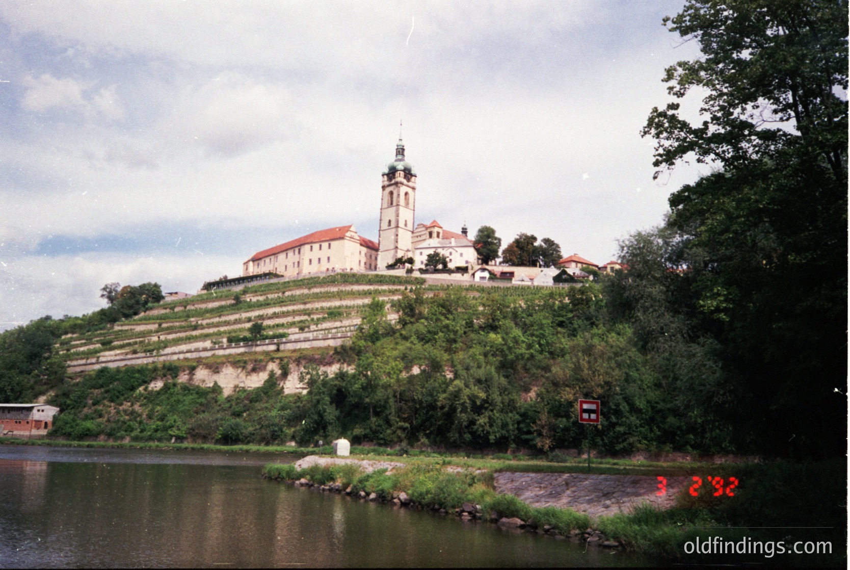 Historic hilltop fortress with red-tiled roofs and a central bell tower, surrounded by terraced greenery. Overlooking a calm riverbank with a stone embankment. Likely Eastern European architecture, possibly or early .