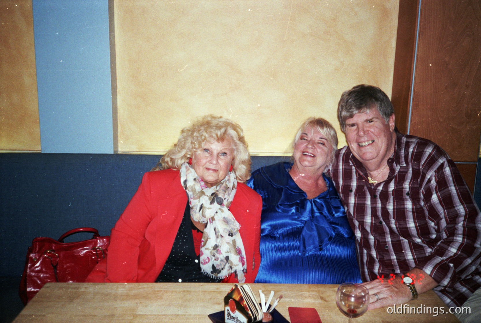 Vintage family portrait featuring three adults in casual indoor setting. Woman on left wears bold red blazer with white fur collar, center wears navy blue dress, man on right sports plaid shirt. Glass table with books and drink holder suggests a relaxed gathering. Warm lighting and muted tones indicate late 20th-century photography.