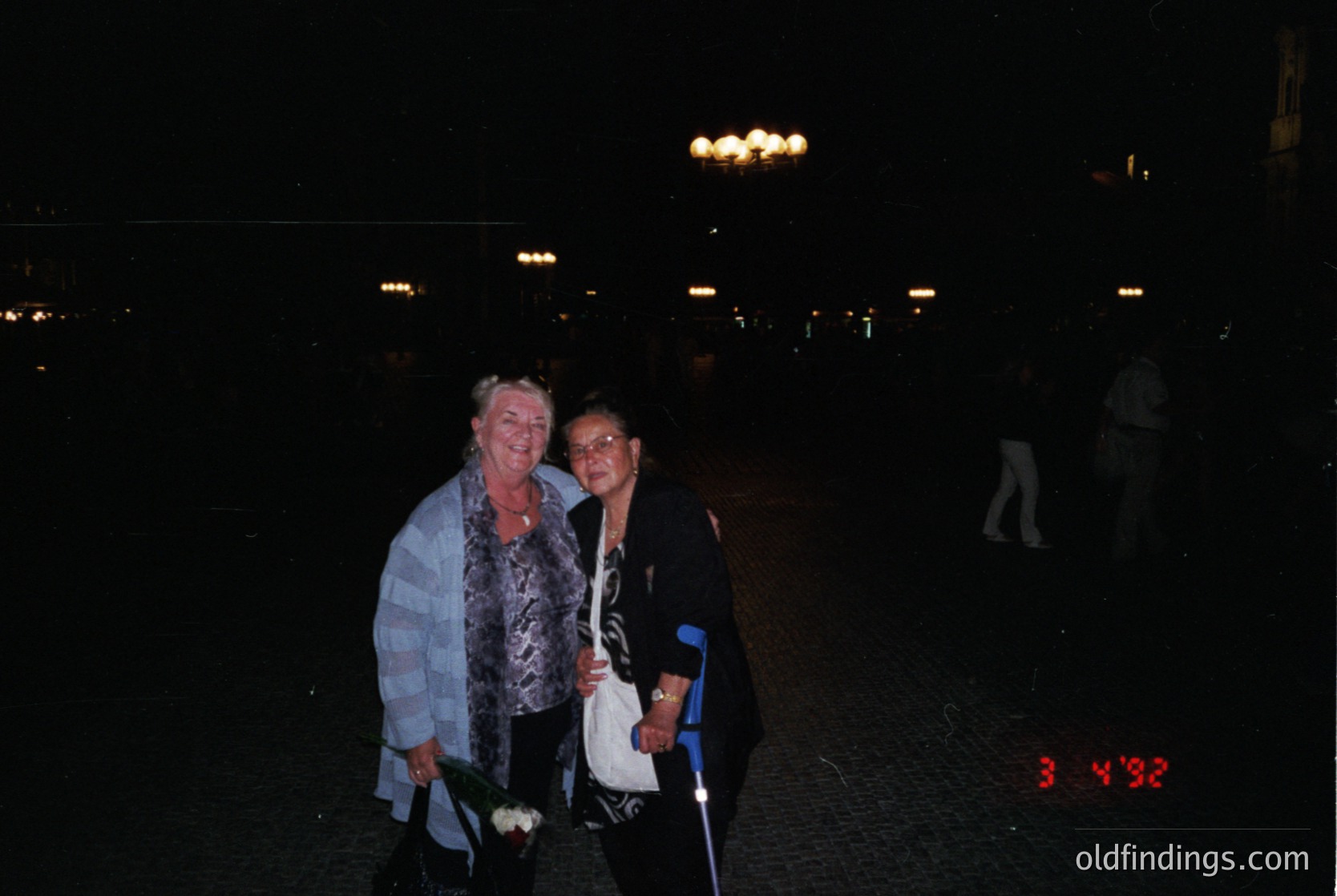 Two women pose under illuminated streetlights in an urban plaza, likely during a nighttime event. The woman on the left wears a light blue, patterned blouse with a checkered scarf, while the woman on the right holds a white clutch and a cane. The timestamp "3:43" suggests late evening. The setting appears modern, possibly mid-to-late 2000s.