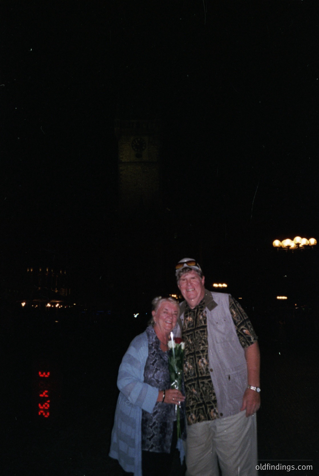 Couple posing under nighttime city lights, holding a single white flower. Man wears patterned shirt, white cap, and light-colored pants; woman in a blue coat. Urban skyline and illuminated signs in background suggest a 1970s-80s setting.