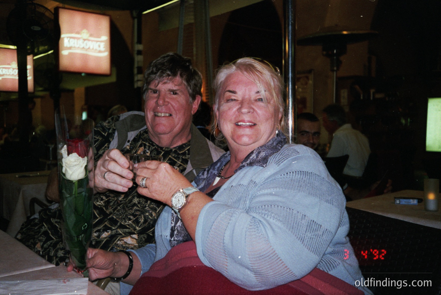 Couple posing indoors at a social event, holding a bouquet of red roses. Man wears patterned shirt, watch, and dark trousers; woman in textured blue sweater. Neon "Kronenbourg" sign and digital clock (30:42) visible. Likely 2000s-era pub or bar setting.