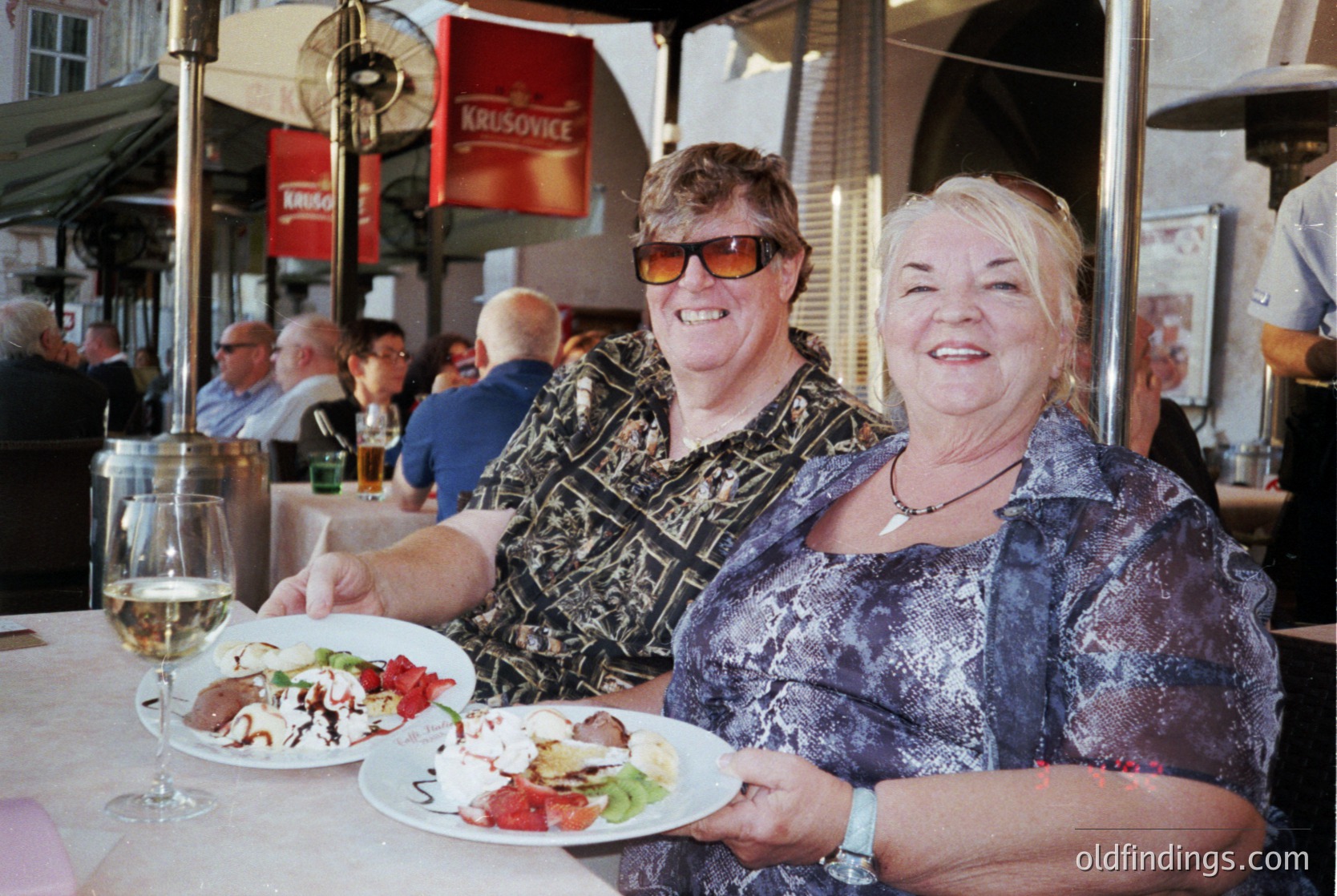 Two women pose joyfully at an outdoor café, holding plates with salads and meats. The woman on the left wears patterned sunglasses and a patterned blouse; the right wears a patterned top and a necklace. Background shows other diners, umbrellas, and signs for "Krusovice" and "Lipton." Likely a European café setting, 2000s–2010s. é