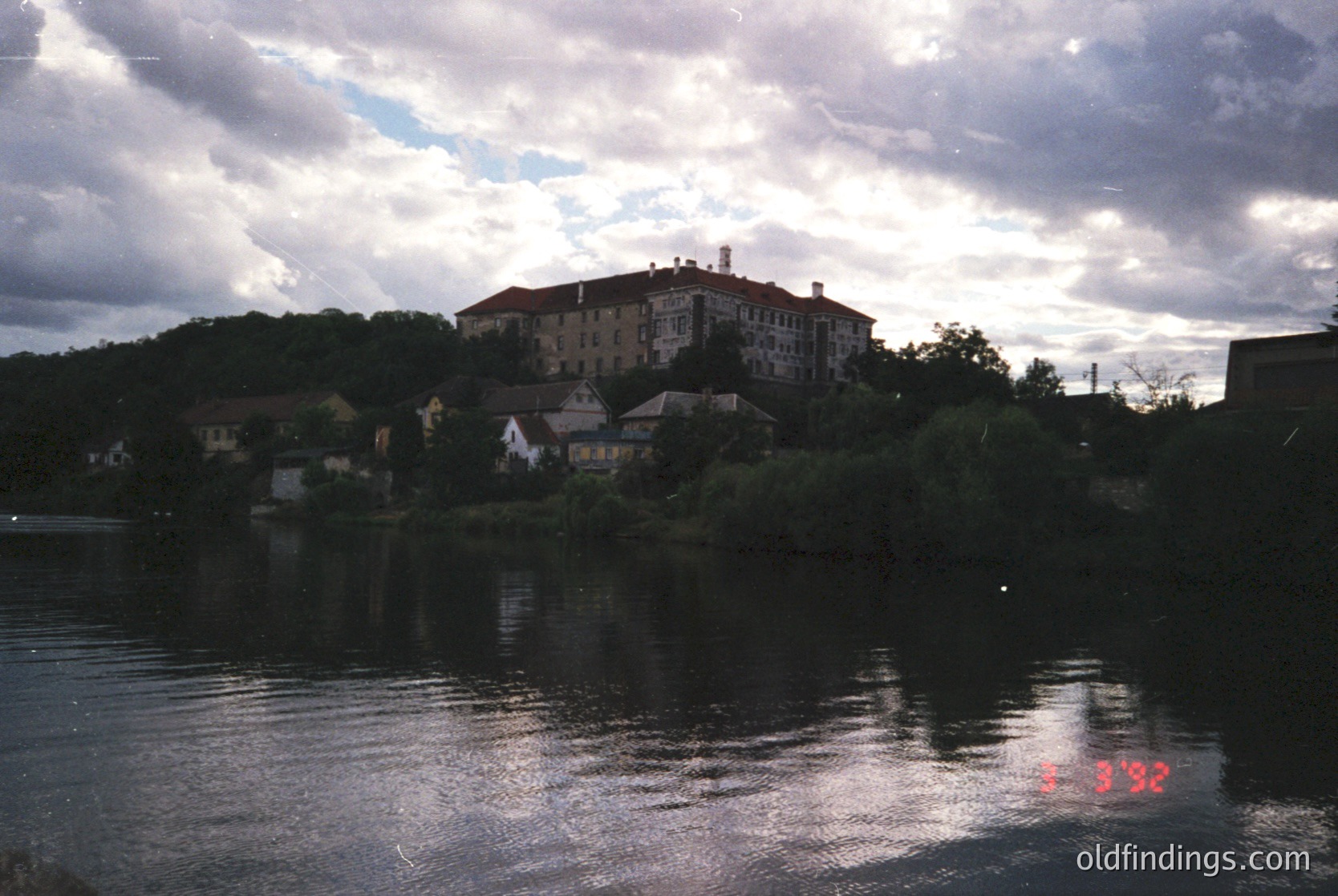 A vintage photograph of a riverside settlement with a prominent multi-story building atop a hill, likely a hotel or institutional structure. Reflections in calm water and dense greenery frame the scene. Overcast skies suggest early/late season lighting. Stamp reads "5.3.92," indicating possible Soviet-era Bulgaria (1992).