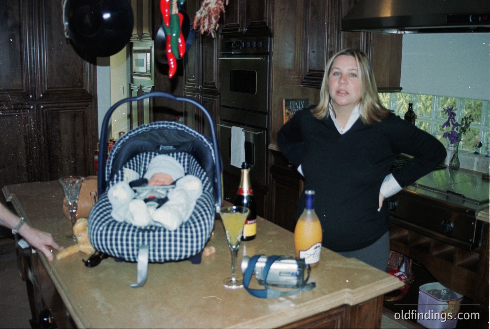 Vintage indoor scene featuring a woman in a 1990s-style sweater posing beside a baby in a checkered carrier on a kitchen counter. Wooden cabinetry, wine bottles, and festive garlands suggest a holiday celebration. Warm lighting and casual decor evoke a mid-1990s to early 2000s domestic setting.