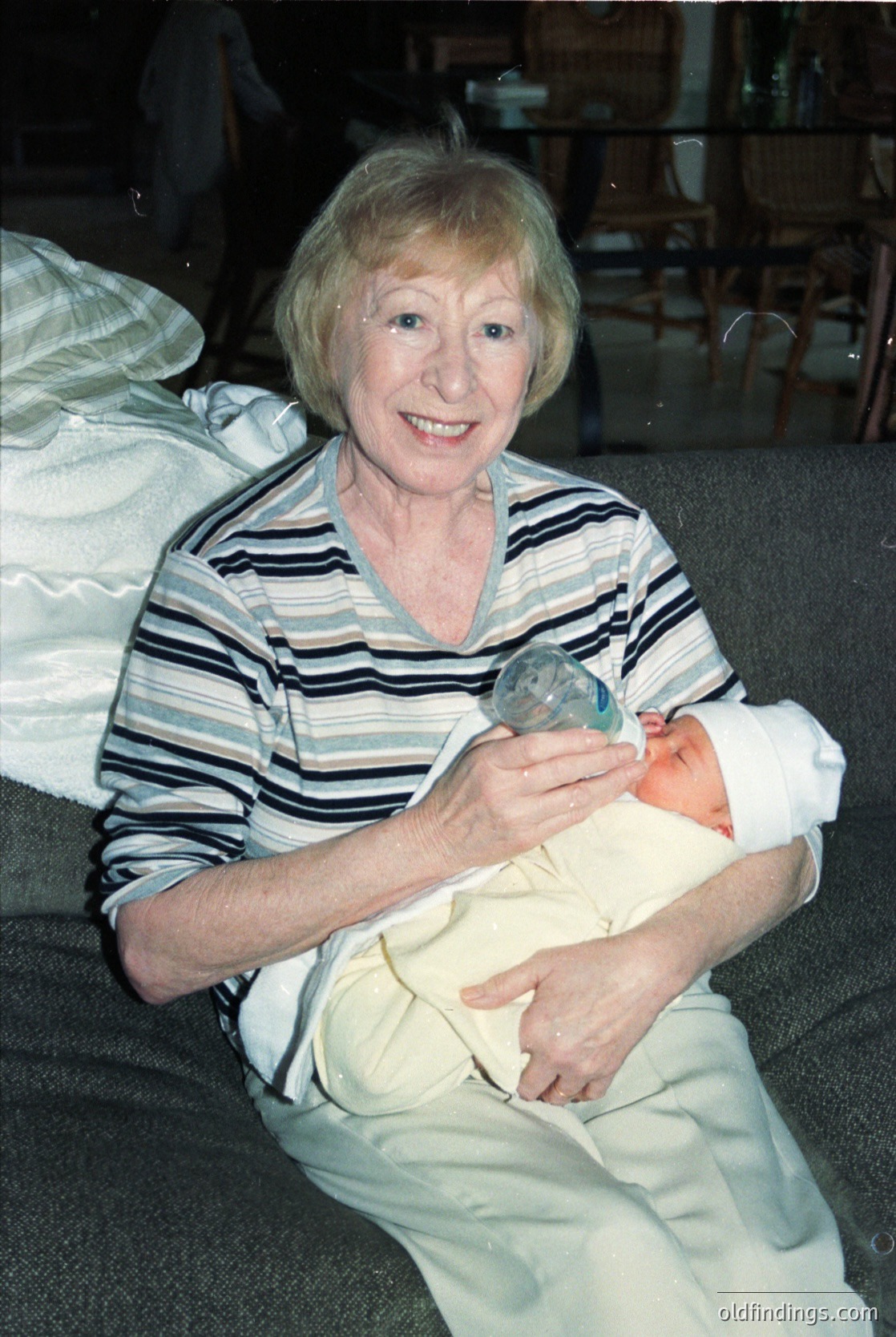 Mid-20th century portrait of an elderly woman cradling a newborn, likely from the 1960s–1970s. She wears a striped blouse and light trousers, holding a glass baby bottle. Background suggests a clinical or hospital setting with medical equipment and a curtained bed.