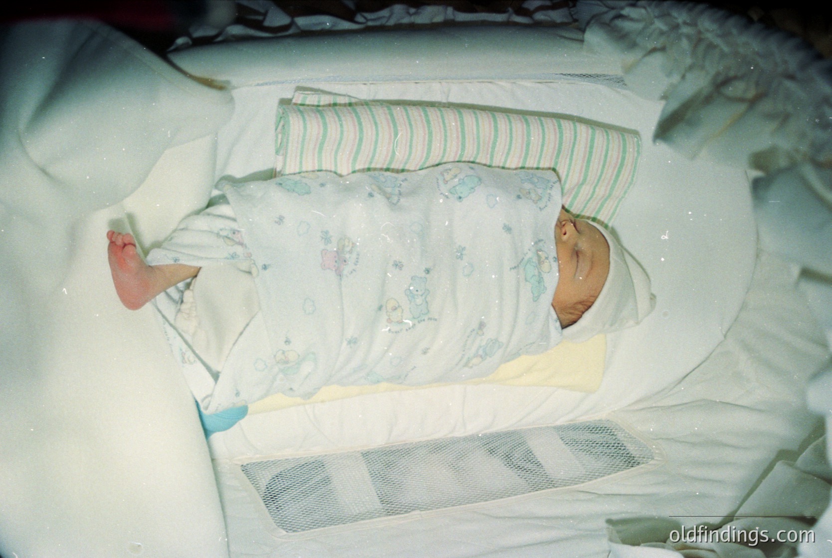 Close-up of an infant swaddled in a vintage floral-patterned onesie, lying on a hospital bassinet with a mesh side panel. Neon lighting suggests a 1980s–1990s hospital setting.