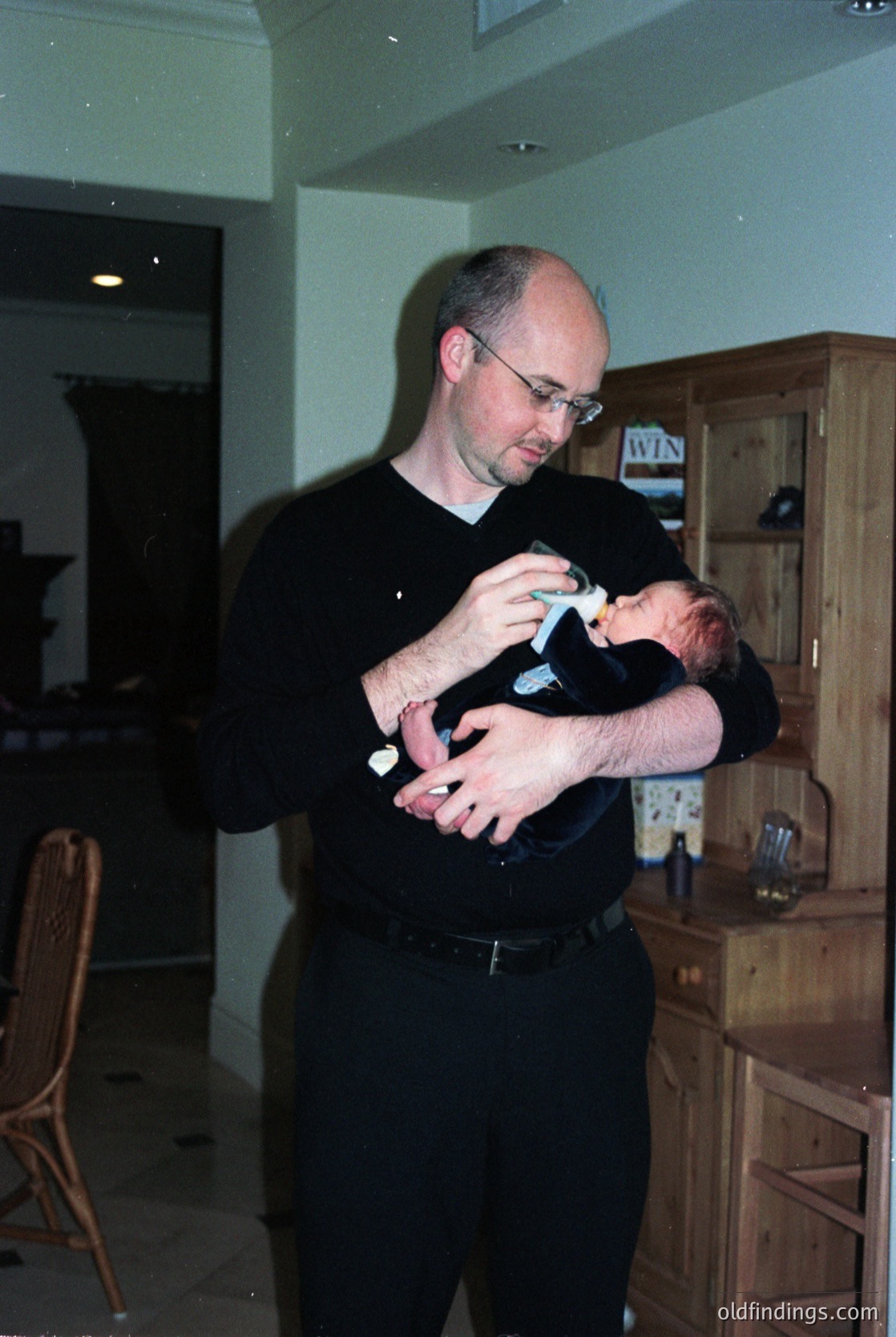 A man in a black sweater cradles an infant in a modern indoor setting, likely a kitchen or living area. Wooden cabinetry with glass-fronted doors and a "WIN" sign on one door suggest a contemporary interior. The scene evokes a candid moment of fatherhood in a residential space.