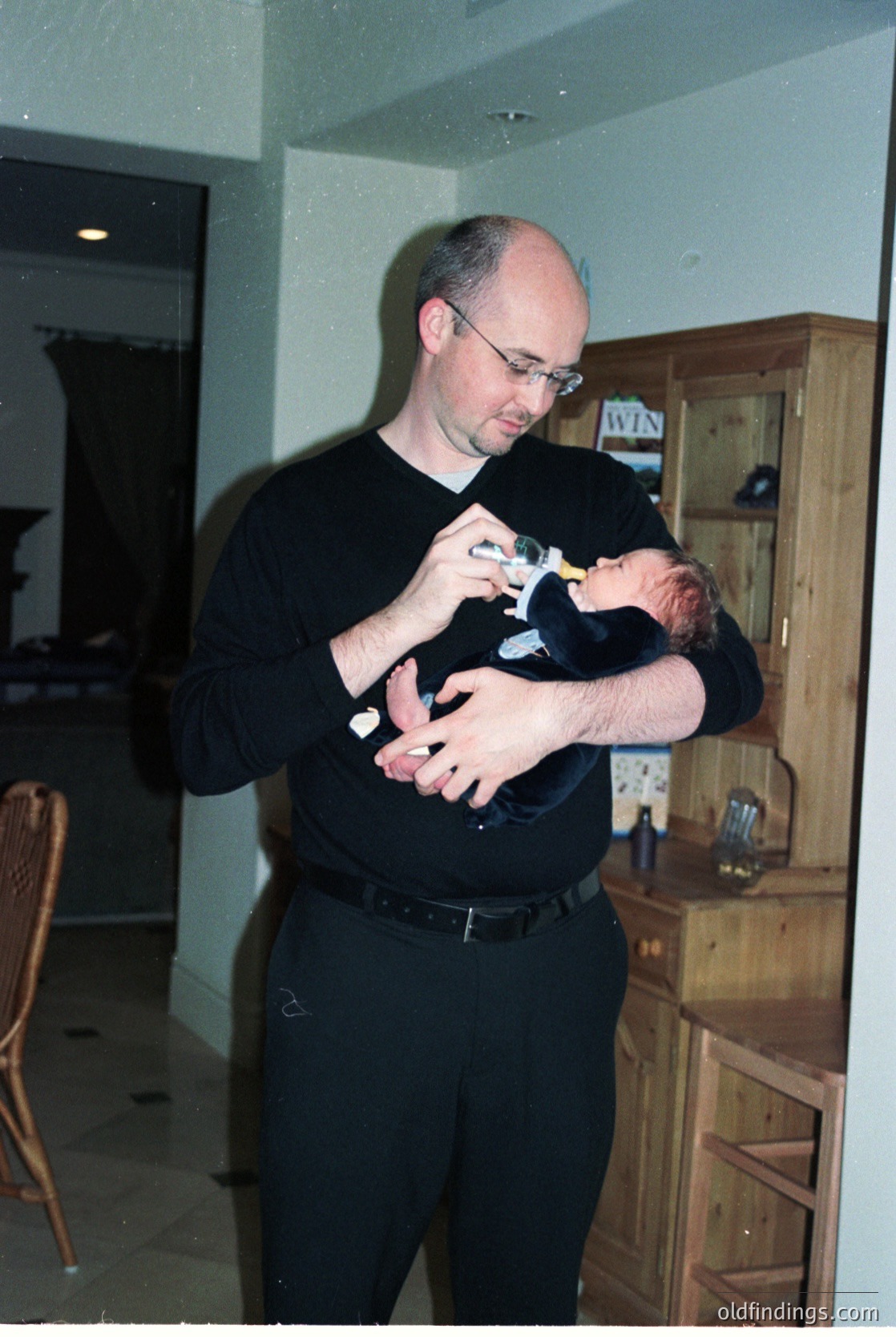 A man in a dark long-sleeve shirt cradles a swaddled infant indoors, likely in a mid-2000s home setting. Wooden cabinetry and a visible "WIN" sticker on the cabinet suggest a domestic kitchen or dining area. The scene captures a tender moment of fatherhood.