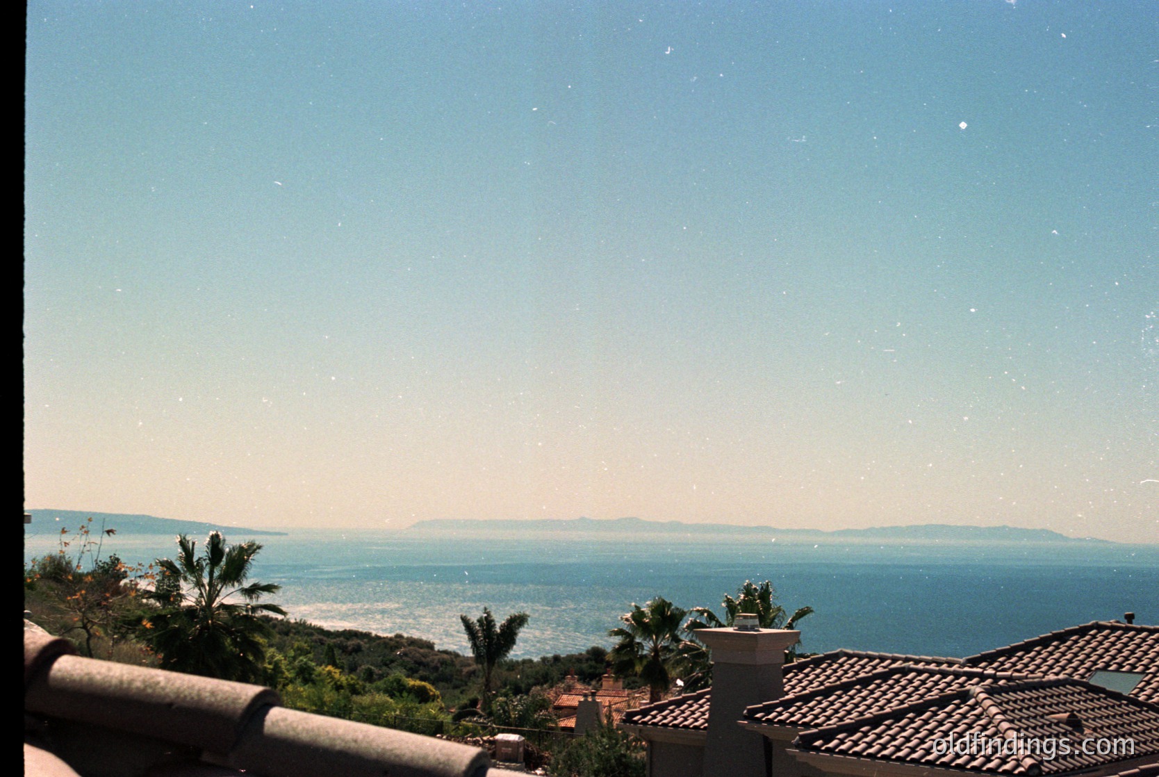 Vintage seaside horizon shot with terracotta rooftops and palm trees in foreground. Clear blue waters meet hazy landmass under star-studded evening sky.