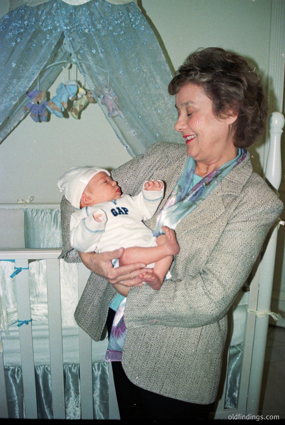 A woman in a 1960s-style sweater cradles a newborn in a hospital bassinet, wearing a diaper with "GAP" branding. Decorative mobiles and a striped curtain frame the scene, suggesting a clinical yet warm nursery setting.