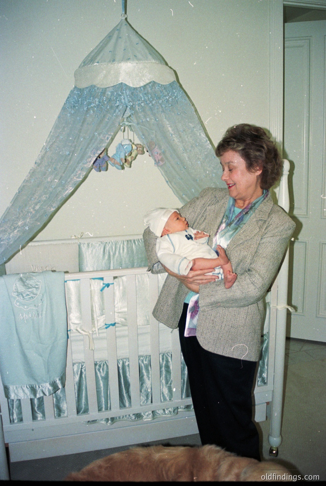 A woman in a 1970s-style cardigan cradles a swaddled infant in a vintage nursery. Decorative canopy bed with blue lace drapes and hanging mobiles frames the scene. Soft pastel tones and classic design evoke mid-century domestic warmth.