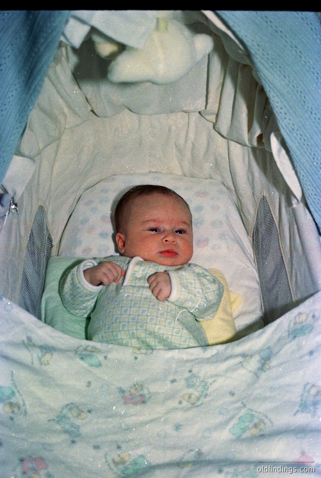 Vintage baby in a white fabric playpen with floral-patterned lining, wearing a crocheted cardigan (green/yellow). Soft focus and warm tones suggest mid-20th century (1950s–1970s) home photography. Playpen design reflects mid-century baby care trends.