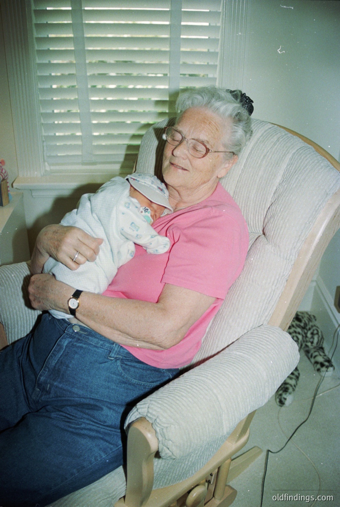 Mid-20th century indoor portrait: Elderly woman in a pink blouse and glasses cradles a swaddled infant in a vintage armchair, surrounded by soft lighting and closed louvered blinds. Ring and watch visible on her wrist. Likely 1950s–1960s, USA/Europe.