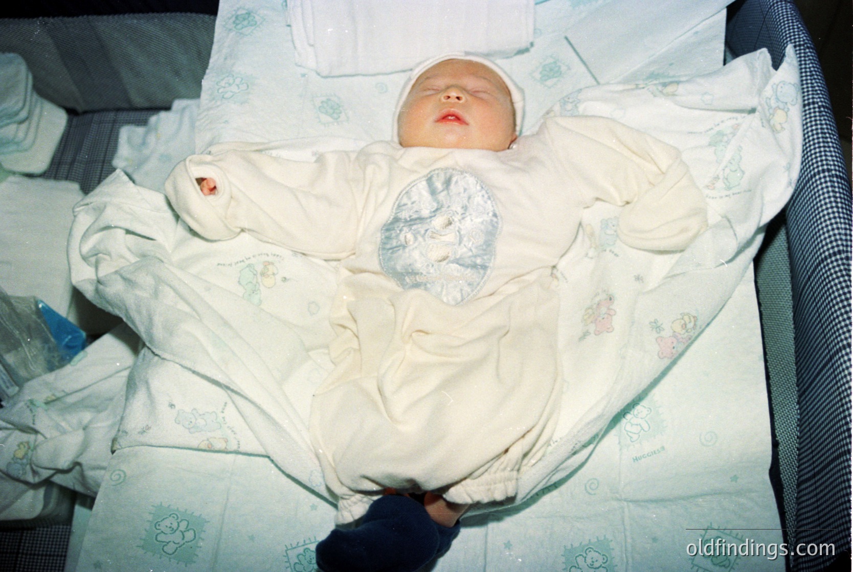 Newborn swaddled in vintage floral-patterned blanket, lying on hospital bassinet with medical tag on chest. Soft focus suggests early 2000s Western hospital setting.