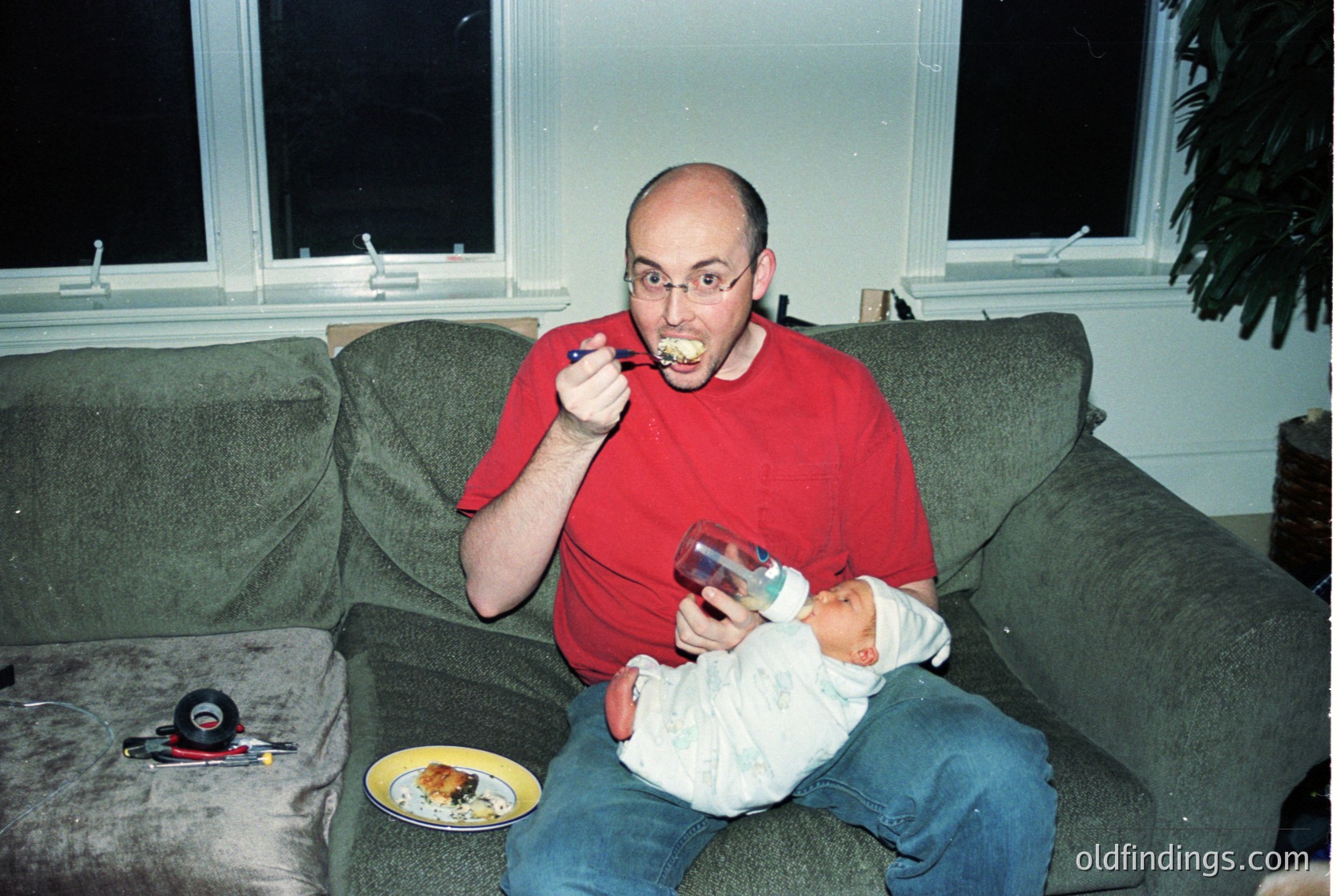 Man in red shirt brushing teeth while holding infant, seated on a vintage-style sofa. Yellow plate with food on armrest. Indoor setting with large window and potted plant. Likely late 1990s–early 2000s domestic scene.