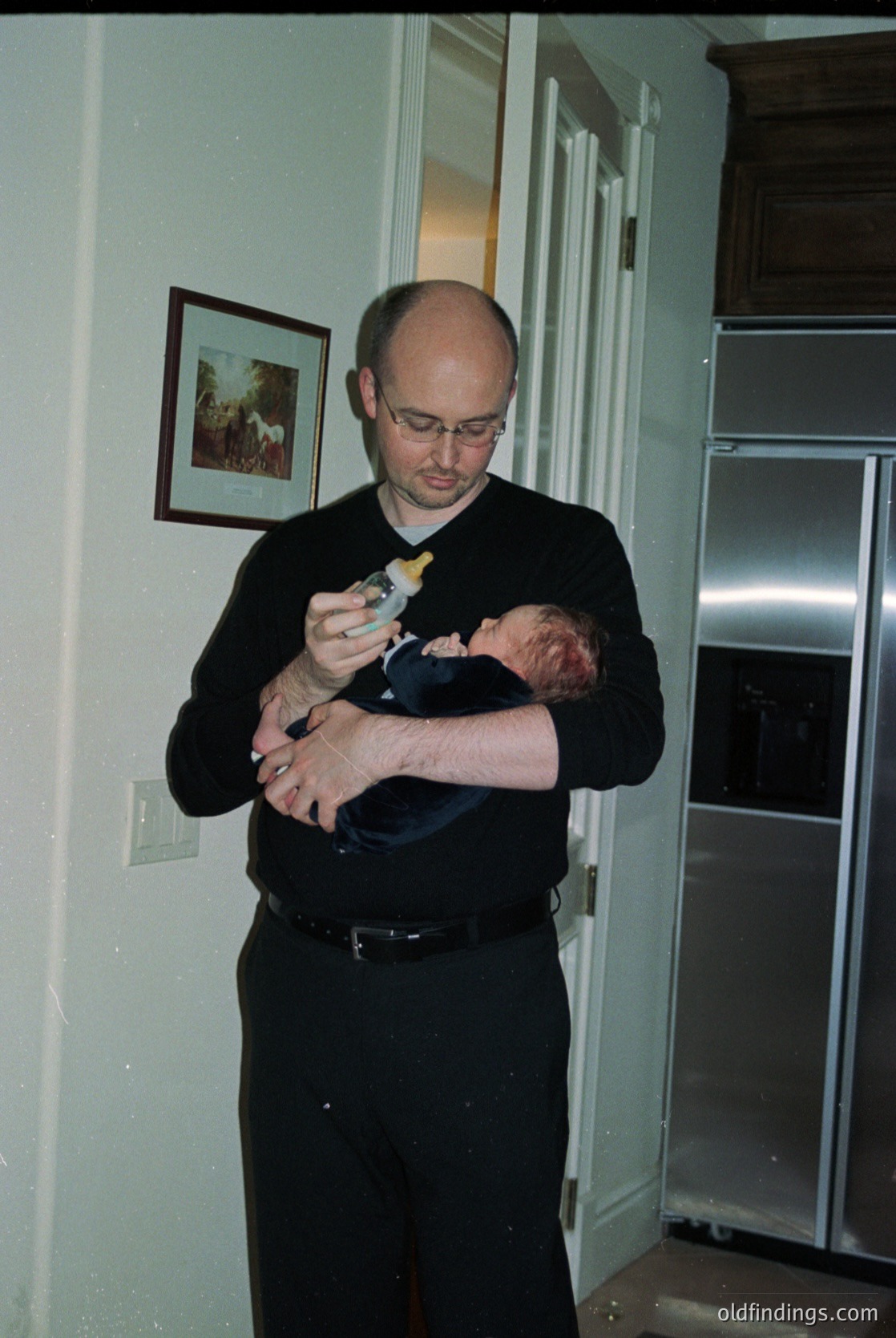 Mid-20th century kitchen interior featuring a man in formal attire (black suit, sweater, glasses) holding a baby while adjusting a toy. Framed artwork on wall, vintage stove, and white paneling. Likely 1960s–1970s domestic setting.
