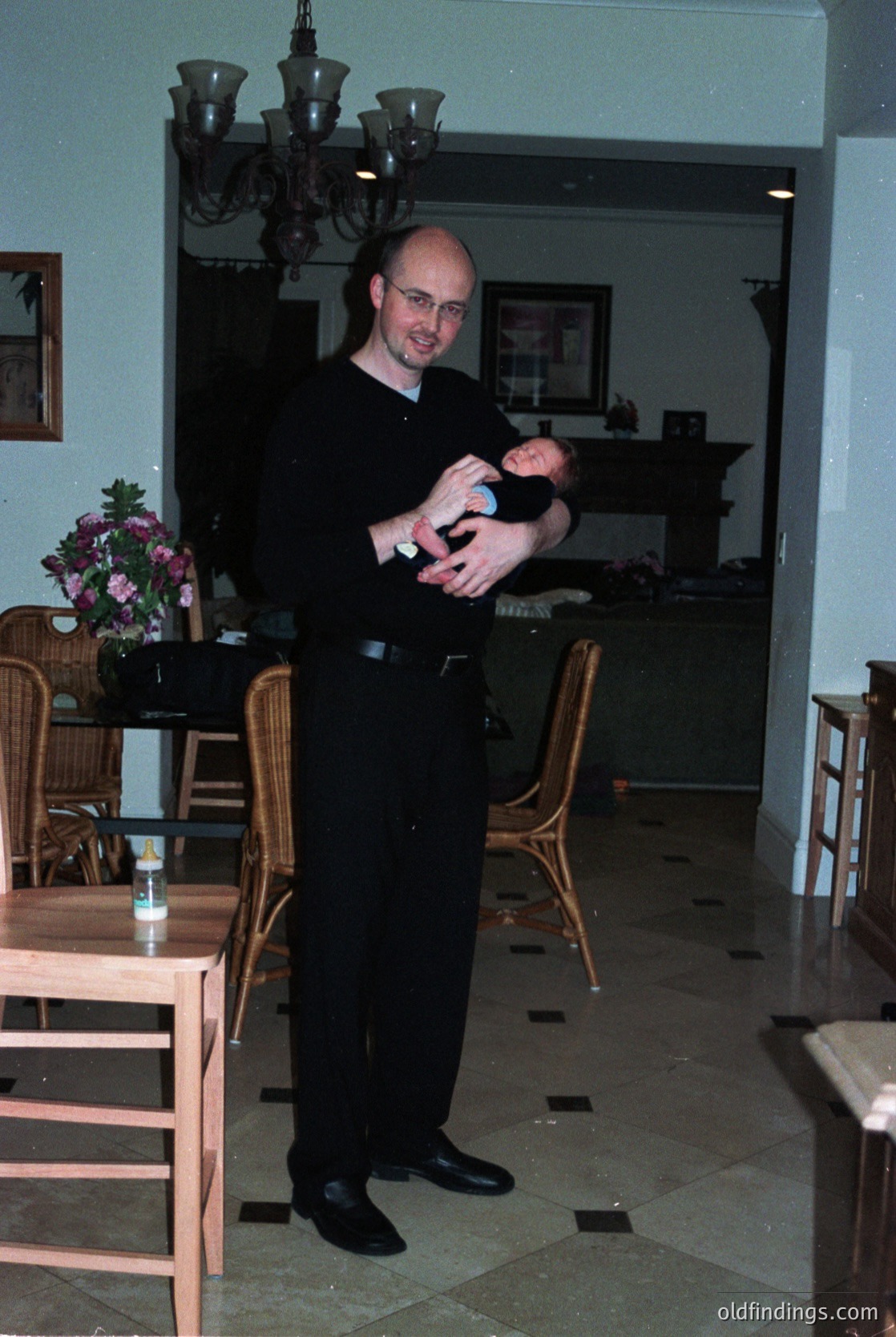 A man in formal attire holds a baby indoors, likely a 1980s-1990s dining/living space. Wooden furniture, floral arrangements, and a chandelier highlight classic decor. Warm lighting and patterned flooring suggest a mid-century European home.
