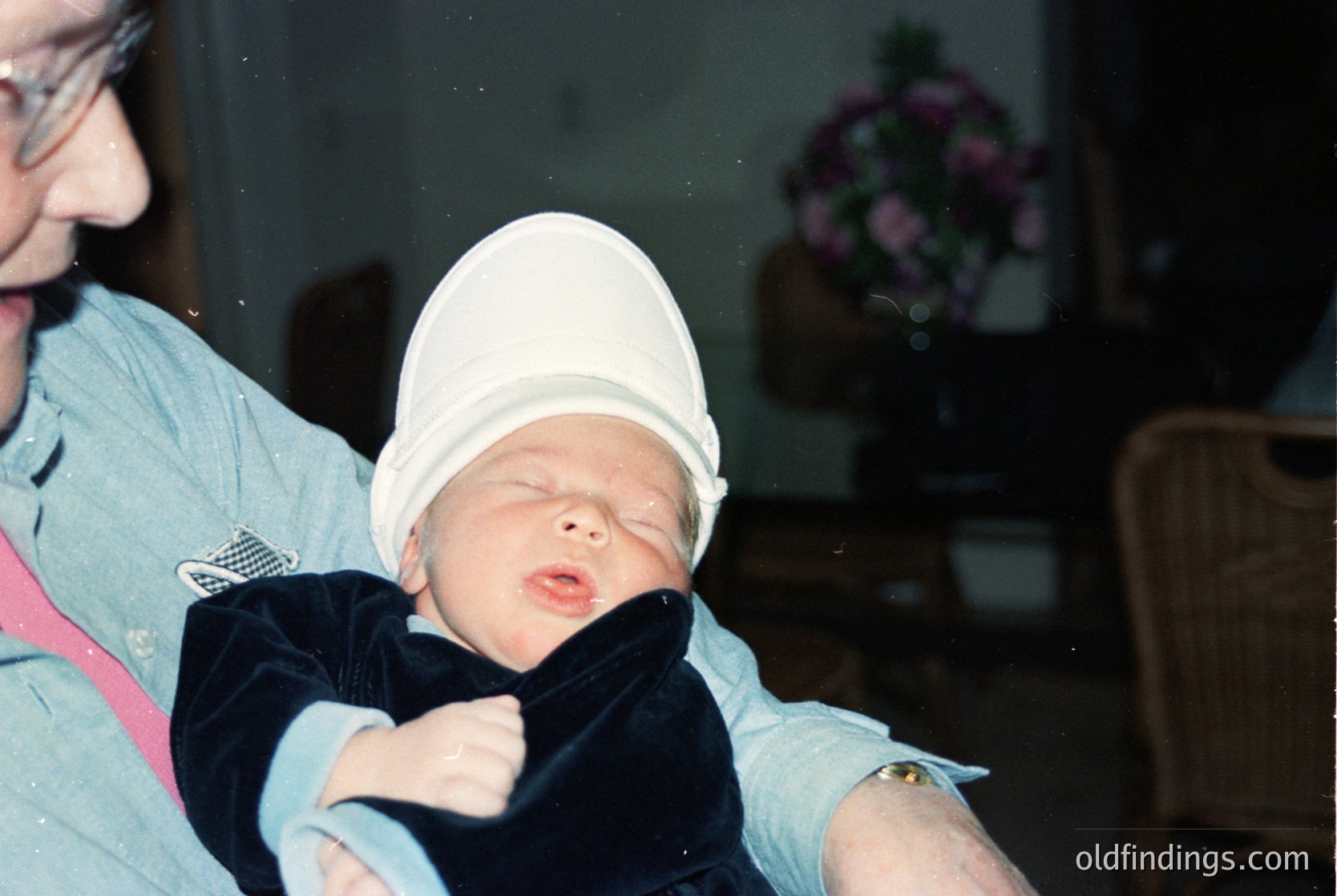 A tender moment captured: an adult holds a newborn in a white knit cap and dark sleeper, indoors near a floral arrangement. Soft lighting suggests a home setting, likely mid-20th century. The image evokes themes of care and infancy.