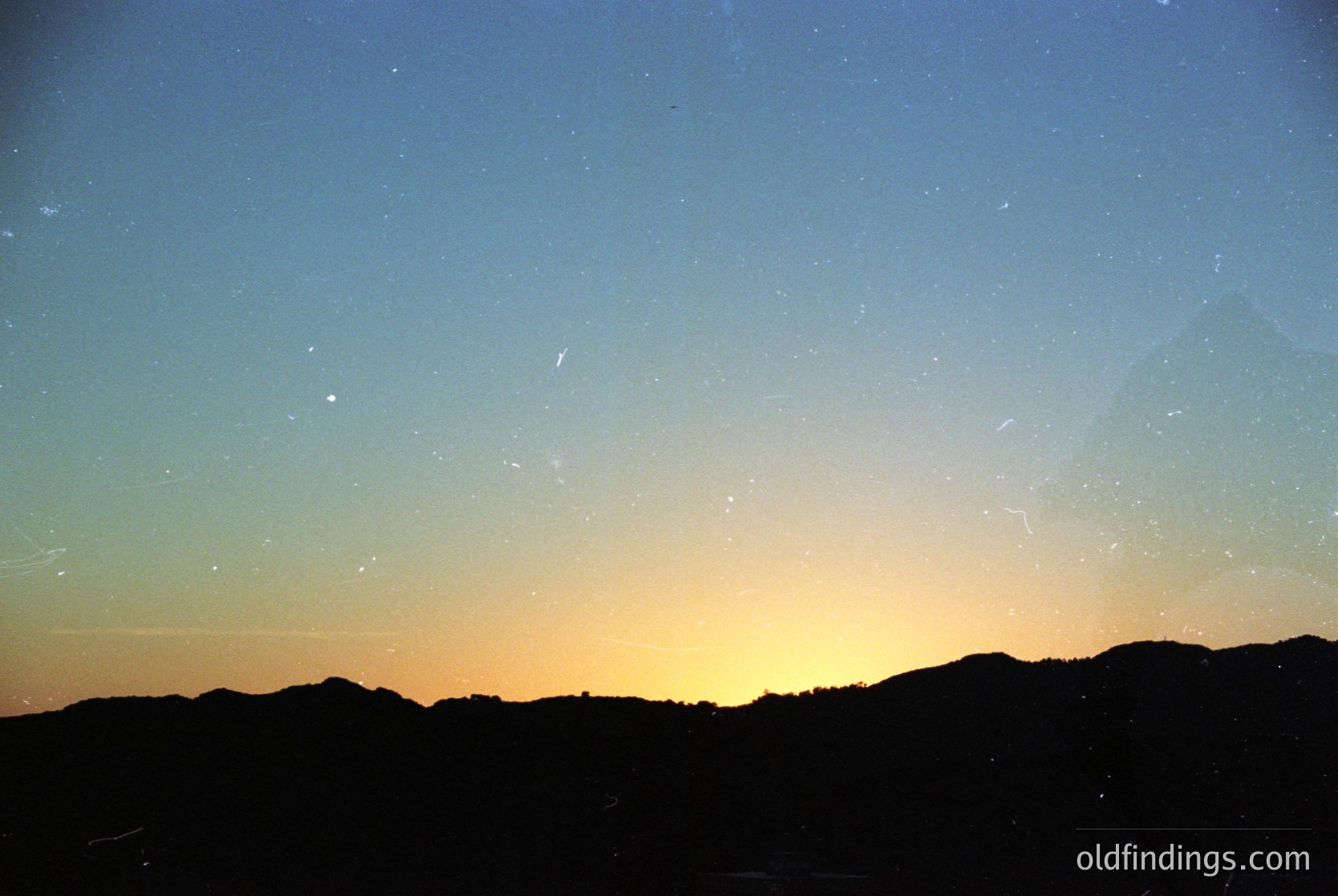 Silhouetted mountain range at dusk with streaks of light pollution and faint meteor trails against a gradient sky (blue to orange). Captures serene natural contrast.