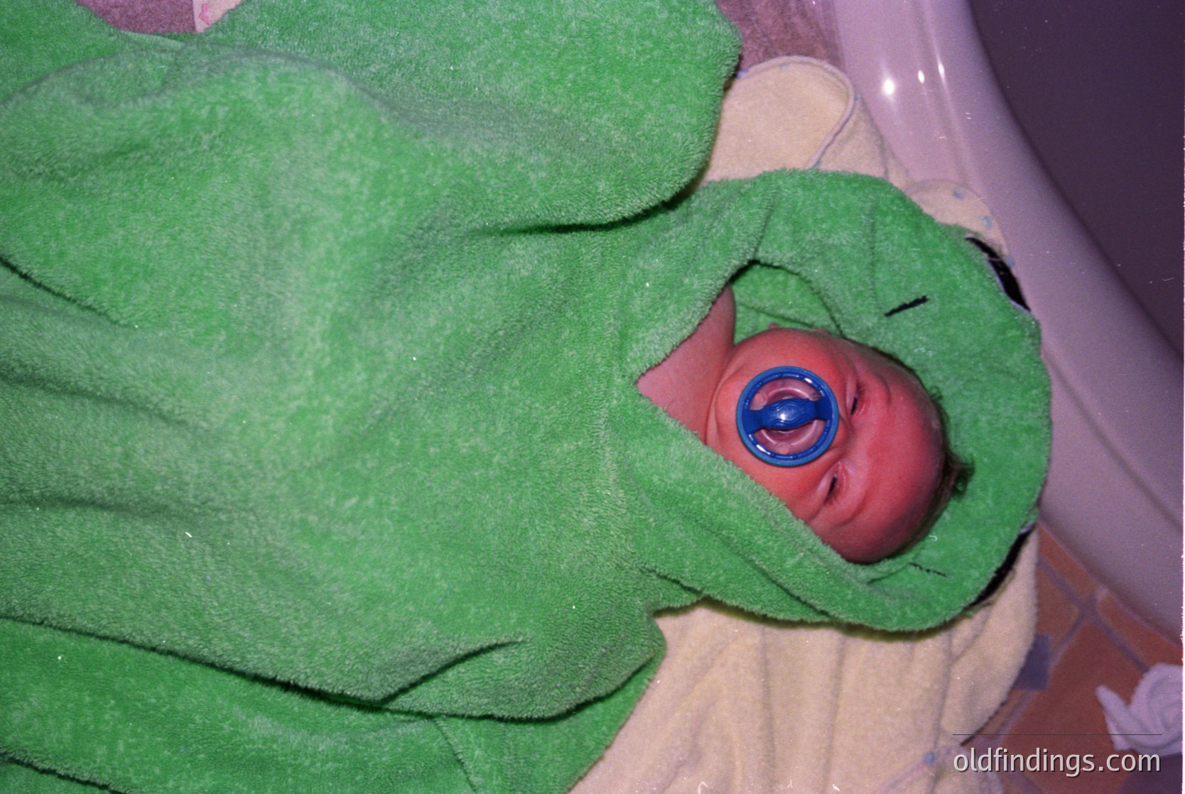 Newborn wrapped in green hospital blanket with blue eye shield, likely for phototherapy. Classic 1970s-1990s neonatal care setup. Warm, clinical atmosphere.