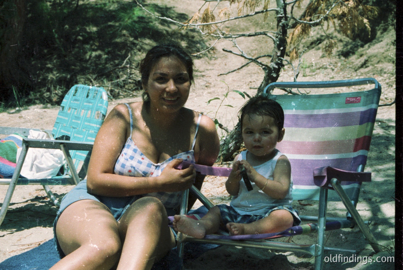 A woman and child relax on vintage beach chairs by a waterfront, likely mid-20th century. The woman holds a pink towel, while the child clutches a toy. Lush greenery and a wooden structure frame the scene.