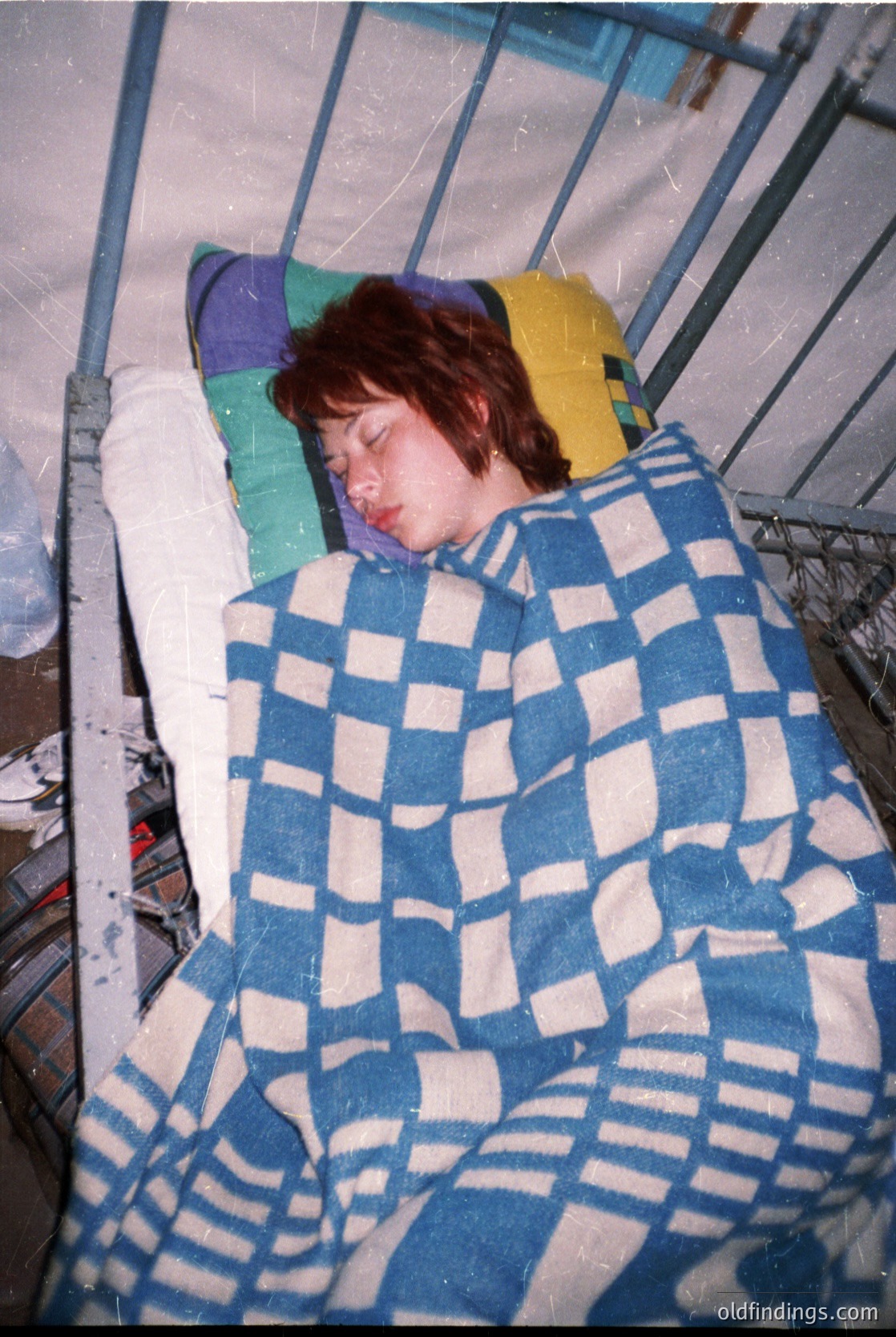 Child resting in a hospital cot, wrapped in a blue-and-white checkered blanket. Medical setting with metal railings and a pillow featuring a yellow and purple design. Likely mid-20th century, suggesting a vintage healthcare aesthetic.