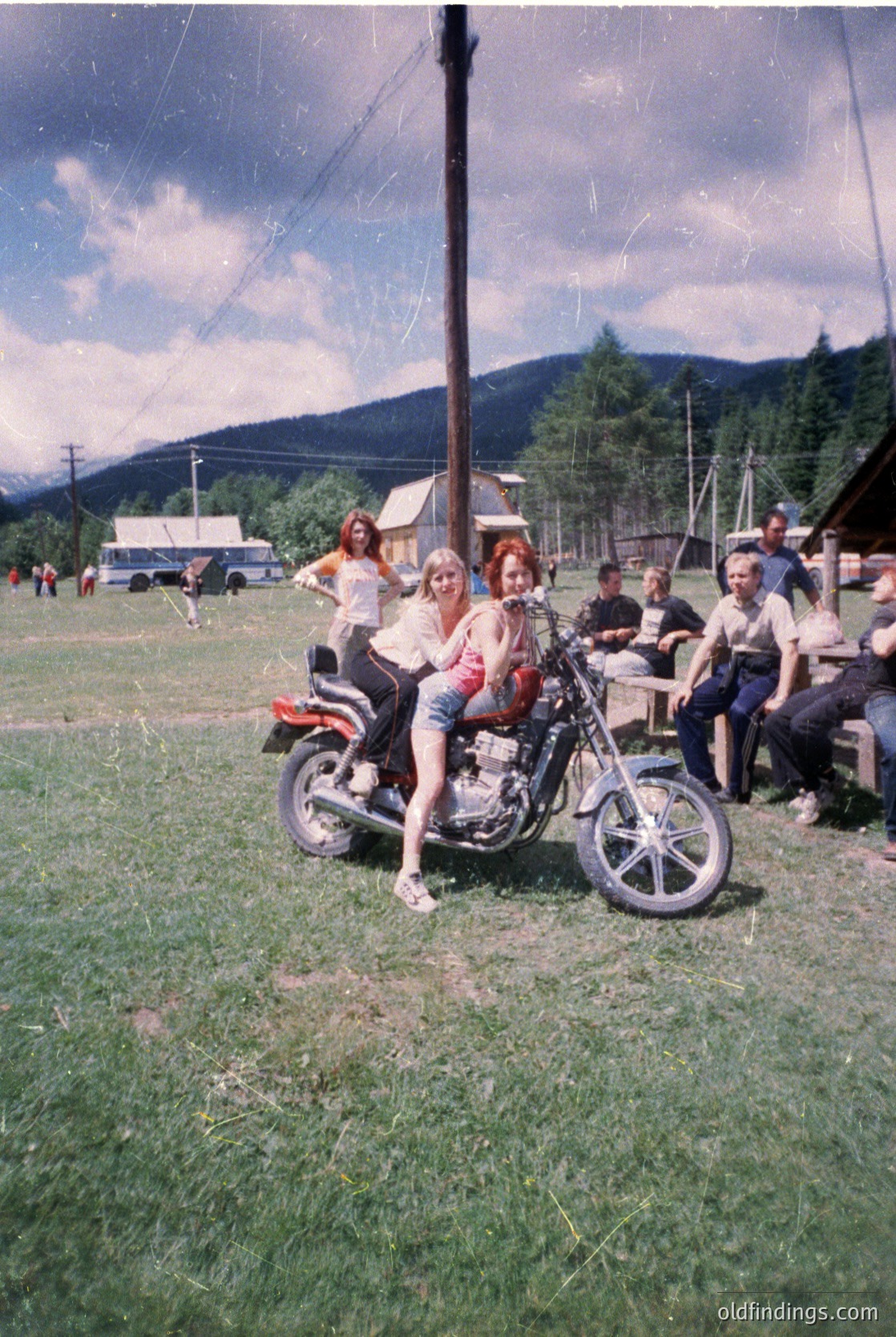 Vintage 1970s outdoor gathering with two women posing on a classic motorcycle in a grassy field. Mountain backdrop and rural setting suggest Eastern Europe. Group of people seated on hay bales, casual summer attire, and cloudy skies enhance nostalgic vibe.