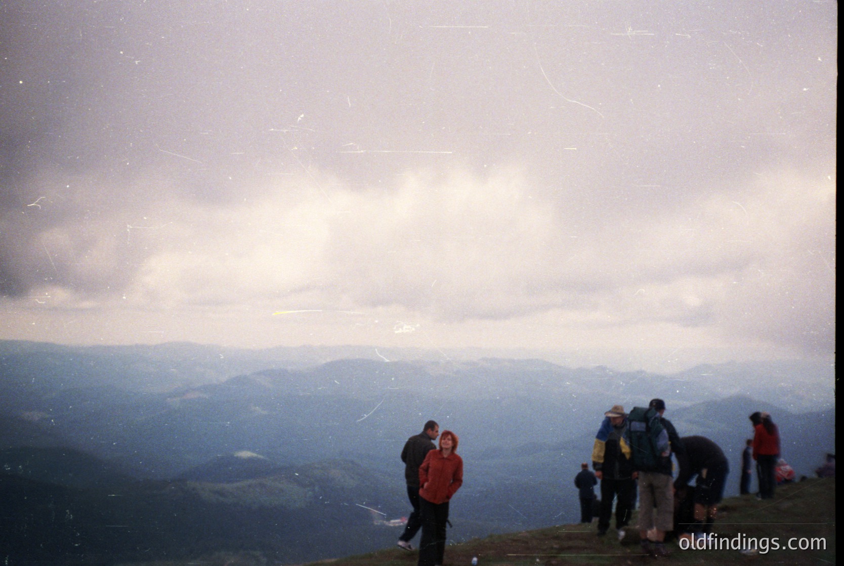 Vintage alpine scene with group of hikers atop a mountain ridge, framed by misty clouds and rolling hills. Distinctive 1980s/90s film grain and sepia tone.