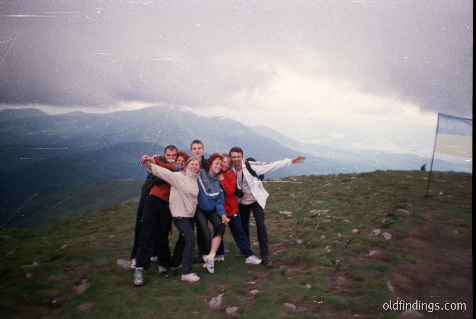 Vintage group photo on a grassy hilltop with mountain backdrop, 1990s-era clothing (tracksuits, sneakers). Five individuals pose with arms around each other, one holding a flagpole. Overcast sky enhances nostalgic, candid vibe.