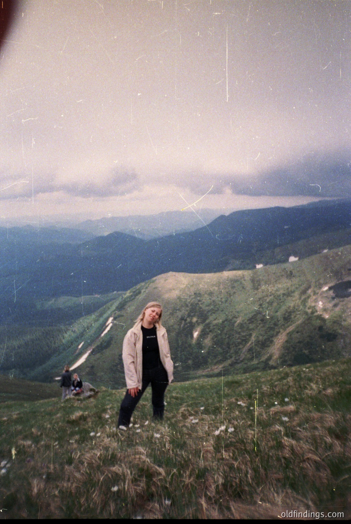 Vintage alpine portrait featuring a lone hiker in mid-20th-century attire (beige jacket, dark pants) standing on a grassy ridge. Dramatic cloud-covered peaks and valleys stretch into the distance, suggesting a remote European mountain range. Film grain and slight fading indicate or era.