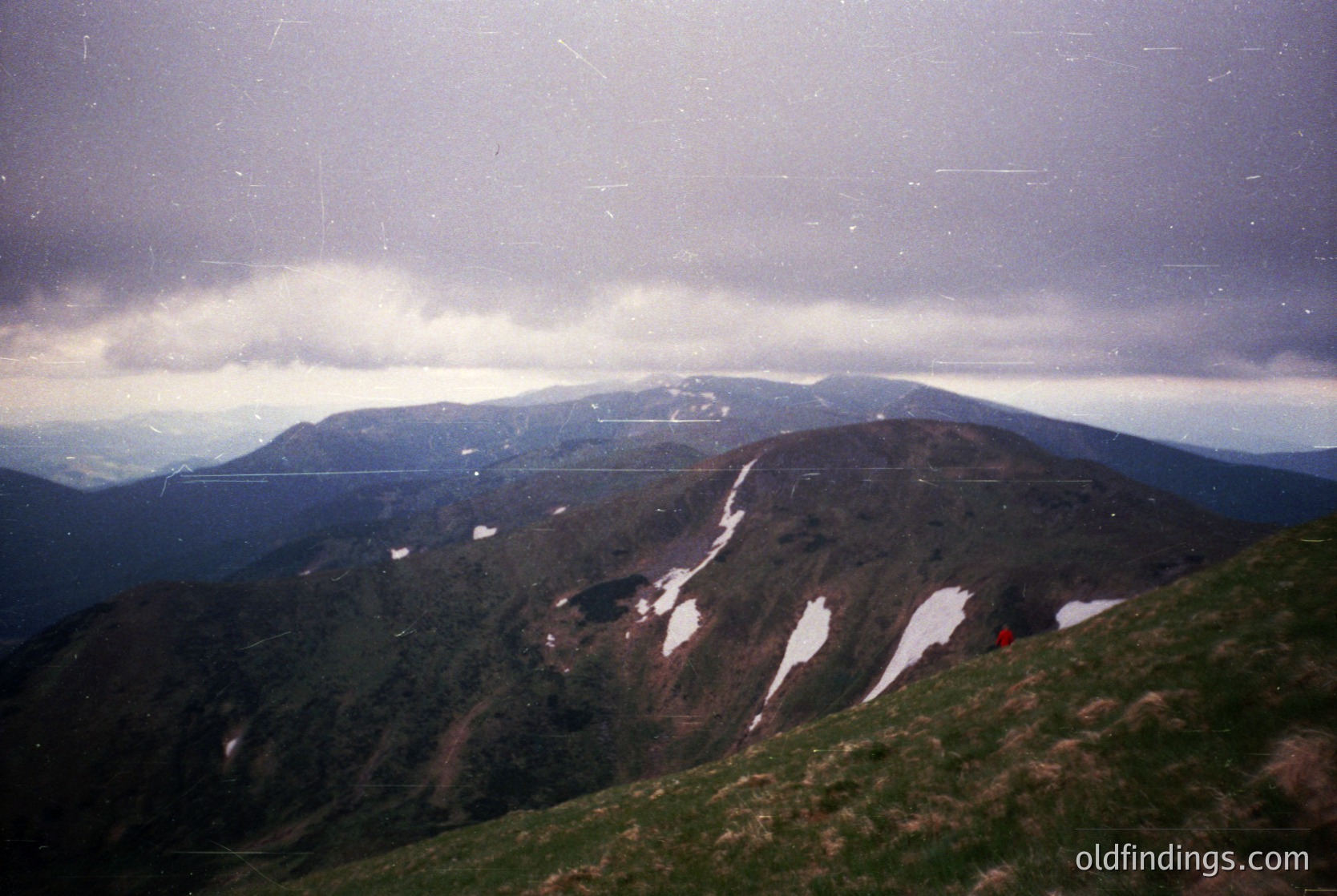 Vintage alpine landscape with snow patches on rocky terrain under low-hanging clouds. Single red backpacker visible mid-hike, suggesting mid-20th century outdoor photography. Dramatic lighting contrasts highlight rugged peaks and misty valleys.