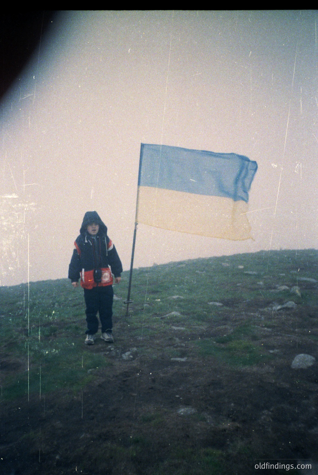 Vintage black-and-white photo of a child holding a Ukrainian flag on a windswept hillside, wearing a red backpack. Distressed film grain and overexposed sky suggest 1960s–1980s Soviet-era photography.