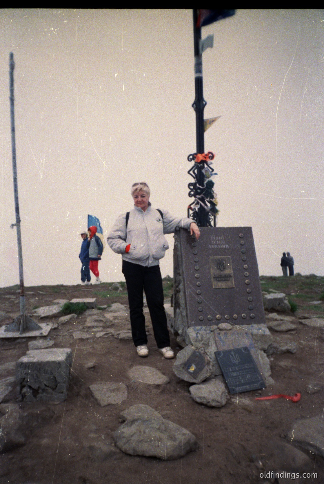 Vintage alpine summit plaque with engraved text ("Vrăciorul" peak, 2508m) and decorative metalwork, set on rocky terrain. Woman in 1970s-style outdoor gear poses beside it. Faded photo suggests mid-20th-century Romanian mountain tourism. ăciorulPeak