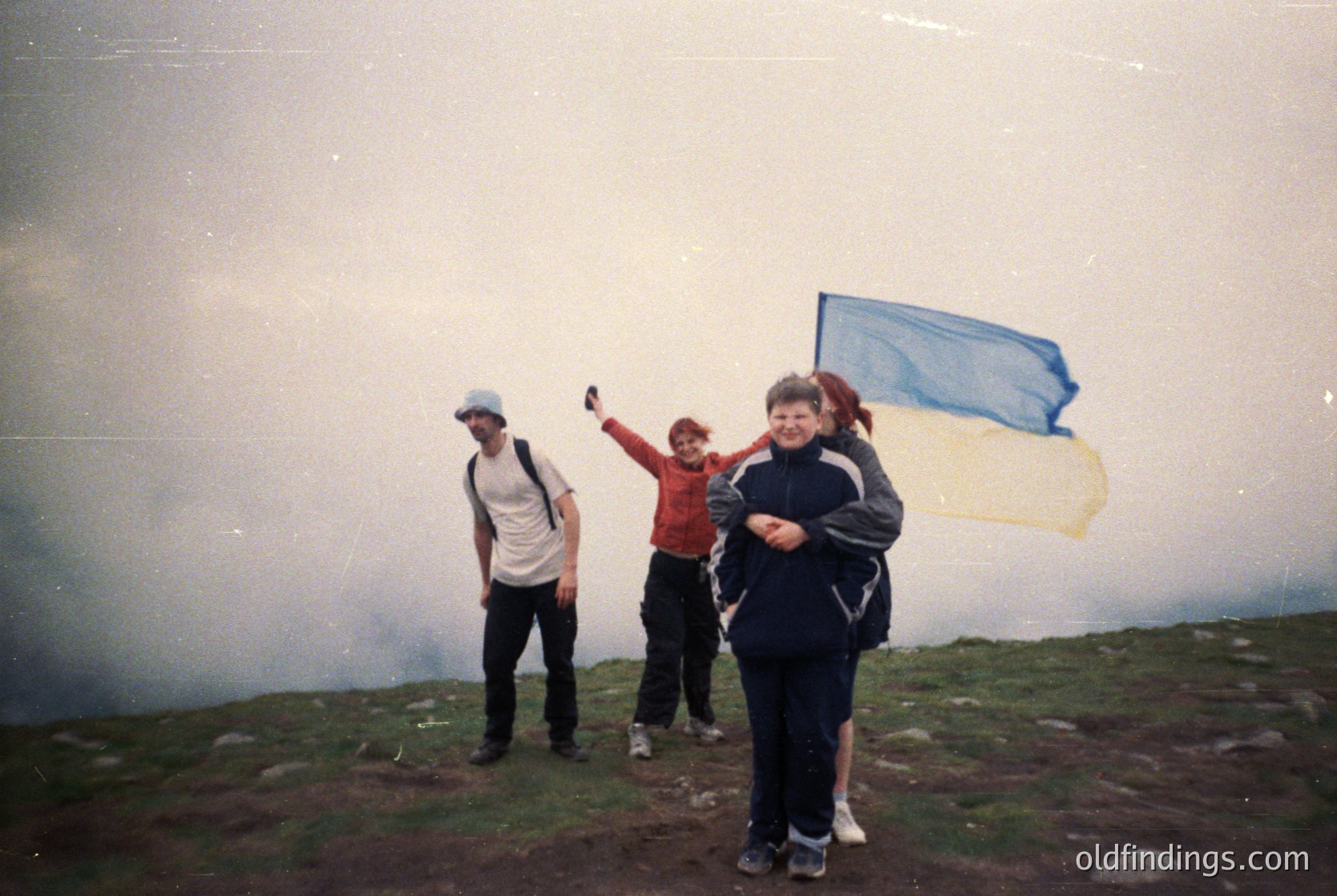 Three hikers pose atop misty mountain peak, 1990s-era gear visible. Blue flag waves in wind, suggesting summit achievement. Fog obscures distant views, highlighting rugged terrain. Casual, outdoor adventure vibe with vintage camera quality.