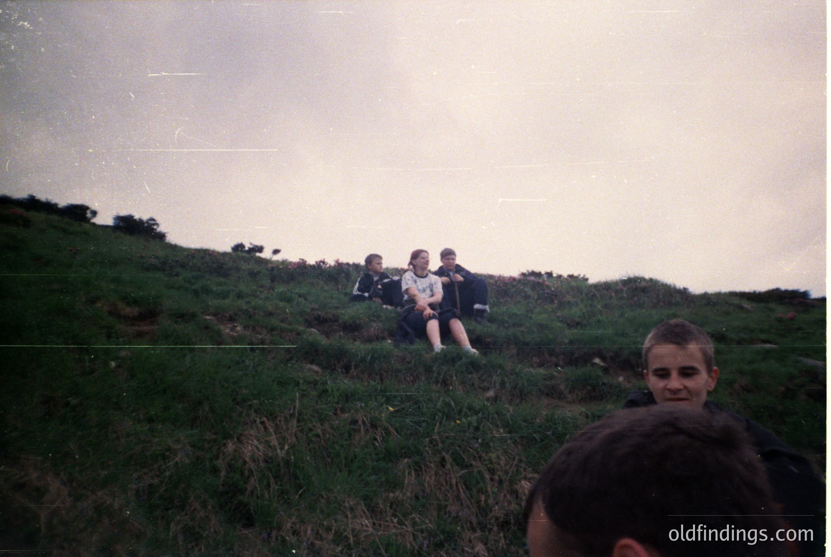 Vintage outdoor group shot on grassy hillside, likely 1980s–1990s. Four seated individuals in matching sportswear (dark jerseys, light shorts) pose casually. Foreground shows a blurred child facing camera. Overcast sky, minimal foliage.