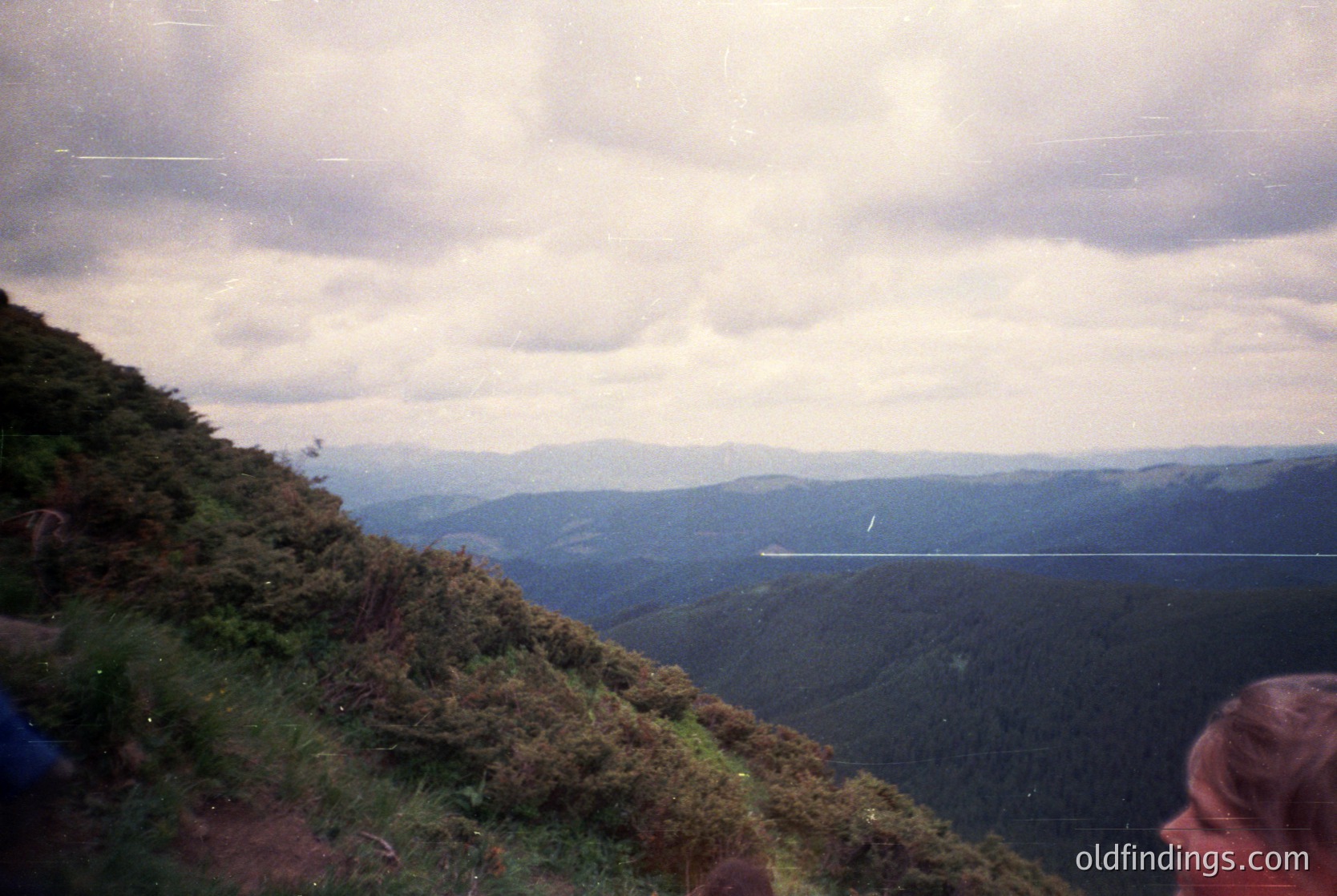 Vintage landscape shot of misty mountain ridges under overcast skies, with a lone road winding through valleys. Foreground shows dense, green vegetation and a blurred figure in profile. Likely 1970s–1990s due to film grain and clothing style.