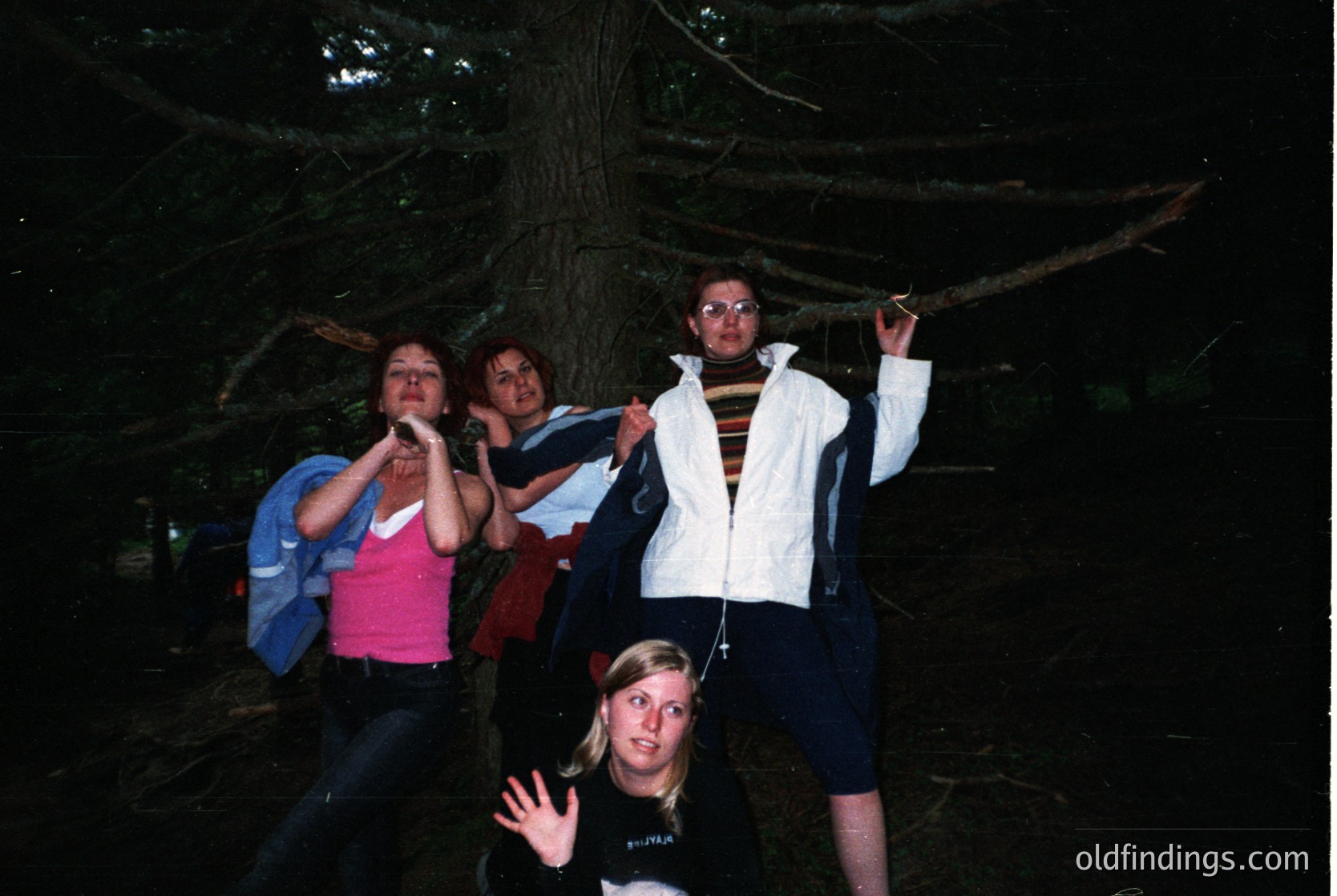 Four individuals pose under a dense forest canopy, likely during a nighttime or low-light hike. Clothing suggests 1990s–2000s casual outdoor fashion: backpacks, layered jackets, and athletic wear. The setting appears to be a temperate forest, possibly North America or Europe. Ideal for stock photography on adventure, travel, or vintage outdoor themes.