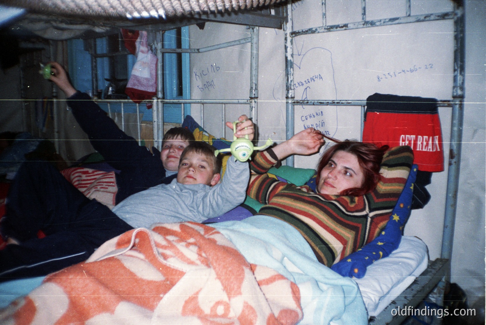 Three individuals pose in a bunk bed dormitory, likely a Soviet-era hostel or barracks. The woman wears a striped sweater and holds a toy, while the child in the middle clutches a stuffed animal. Bunk beds, metal bars, and handwritten Cyrillic notes on walls suggest a utilitarian, shared living space. The red "GET REAL" bag hints at 1990s Western influence.