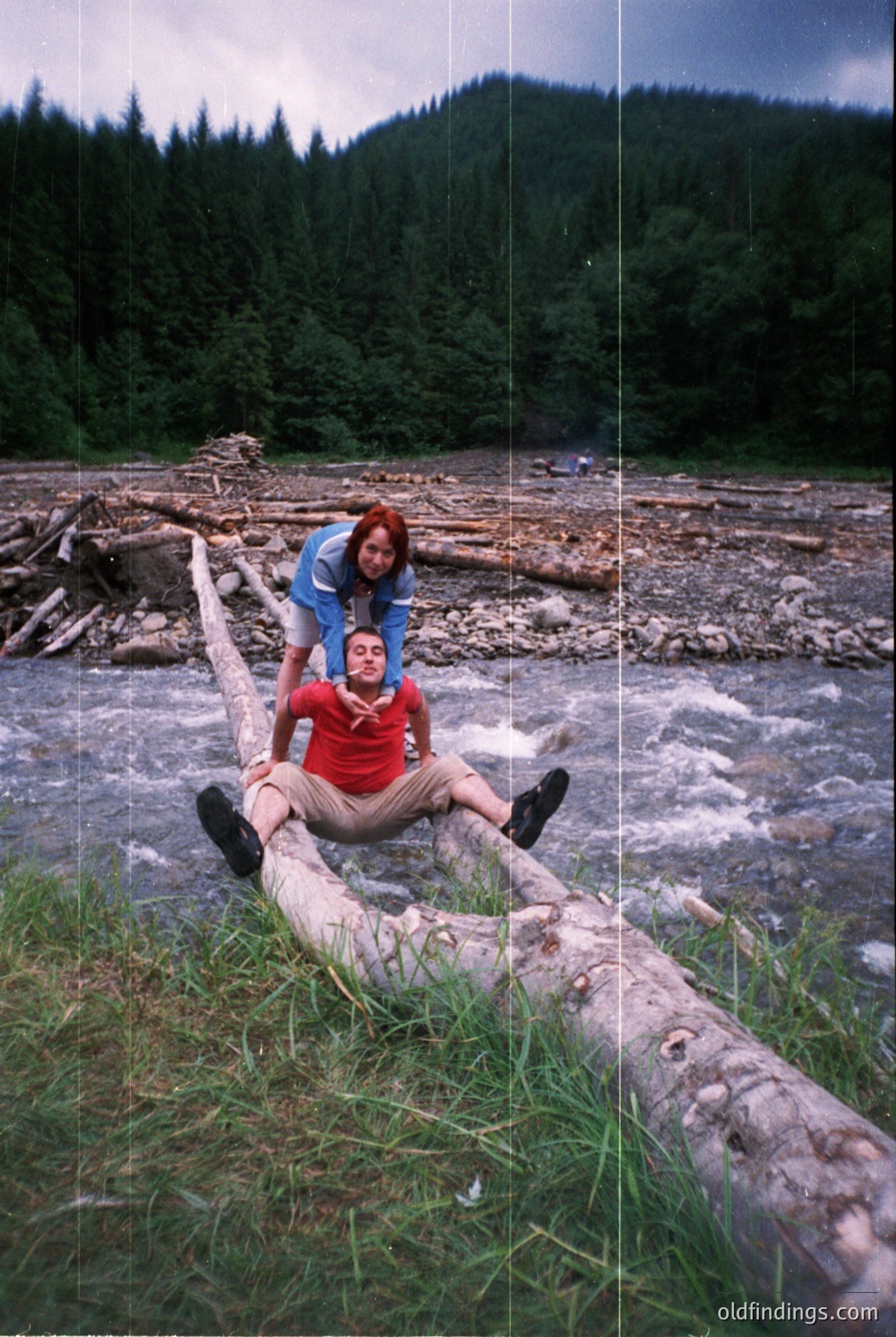 Two individuals pose on a fallen log near a flowing river, surrounded by forested hills. The man wears a red shirt and beige pants, while the woman stands behind him in a blue jacket. The scene suggests a mid-20th-century outdoor adventure, likely in a temperate forest region. [Couple posing on fallen log by river in forested hills, mid-20th century ]