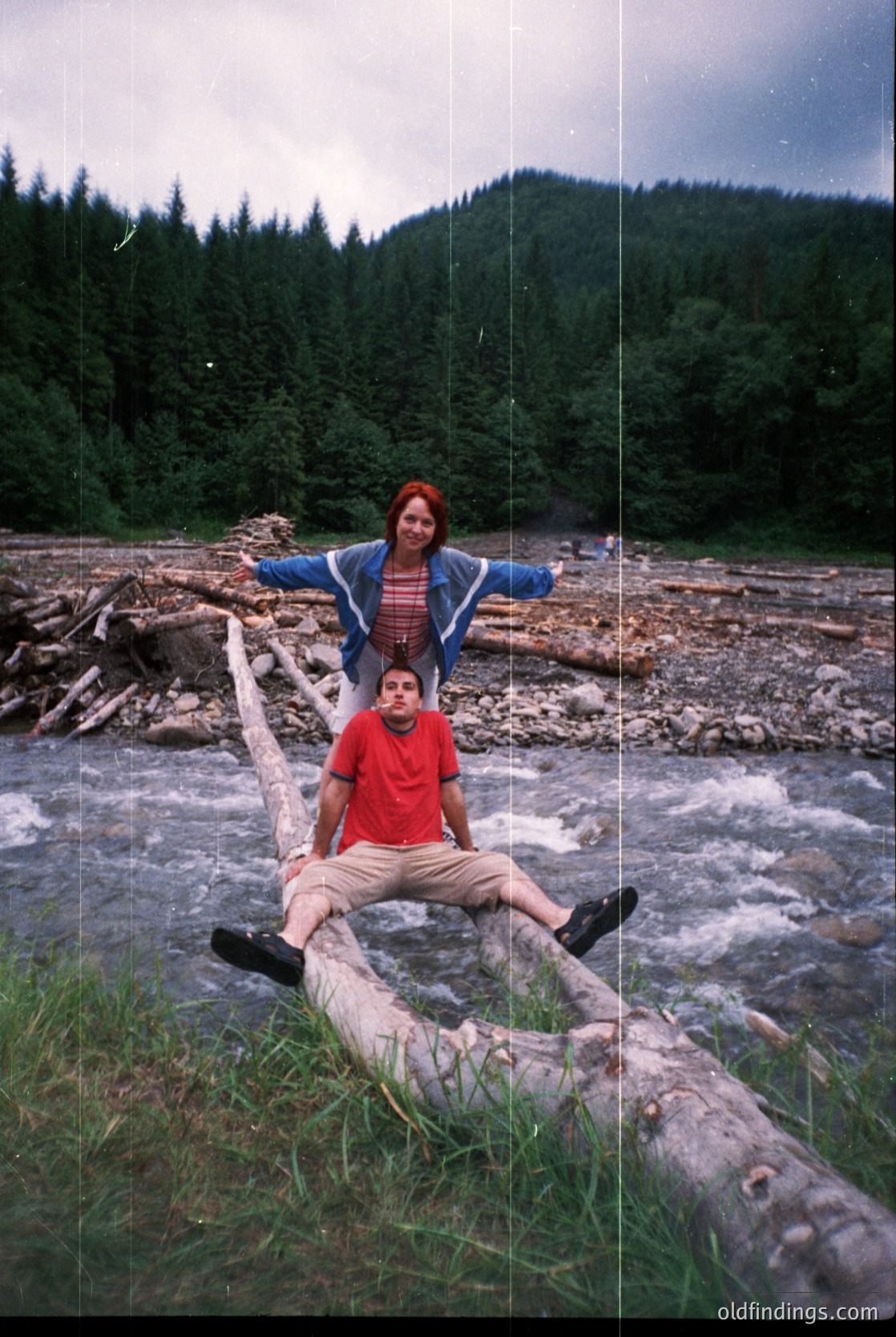 Two individuals pose on a fallen log over a shallow, rocky riverbed, surrounded by dense forest. The woman stands behind the seated man, arms outstretched. Mid-20th century clothing suggests a or setting. Lush greenery and misty mountains hint at a European alpine region. [Family adventure in misty alpine forest, 1960s-70s ]