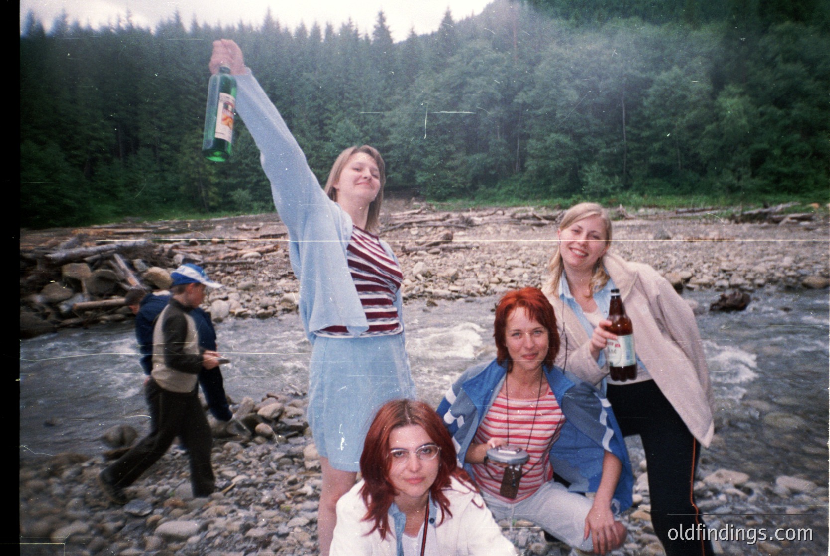Vintage group photo by a rocky riverbank surrounded by dense forest. Four women pose with drinks, mid-1990s Eastern European attire (striped shirts, denim). Background shows a blurred figure walking.