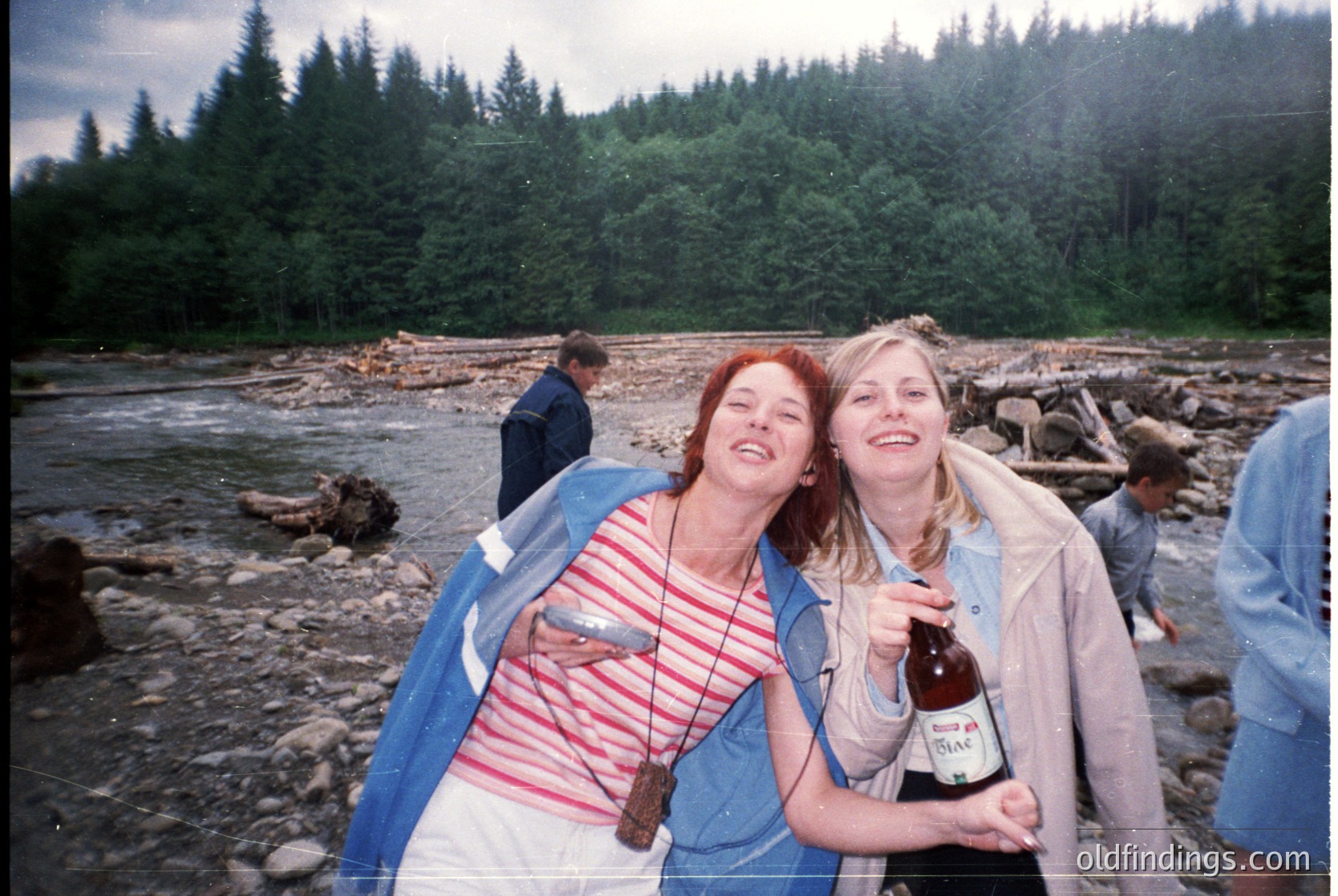 Two women pose joyfully by a rocky riverside in lush forest, holding a bottle of beer. Casual 1990s outdoor gathering with vintage clothing and natural scenery.