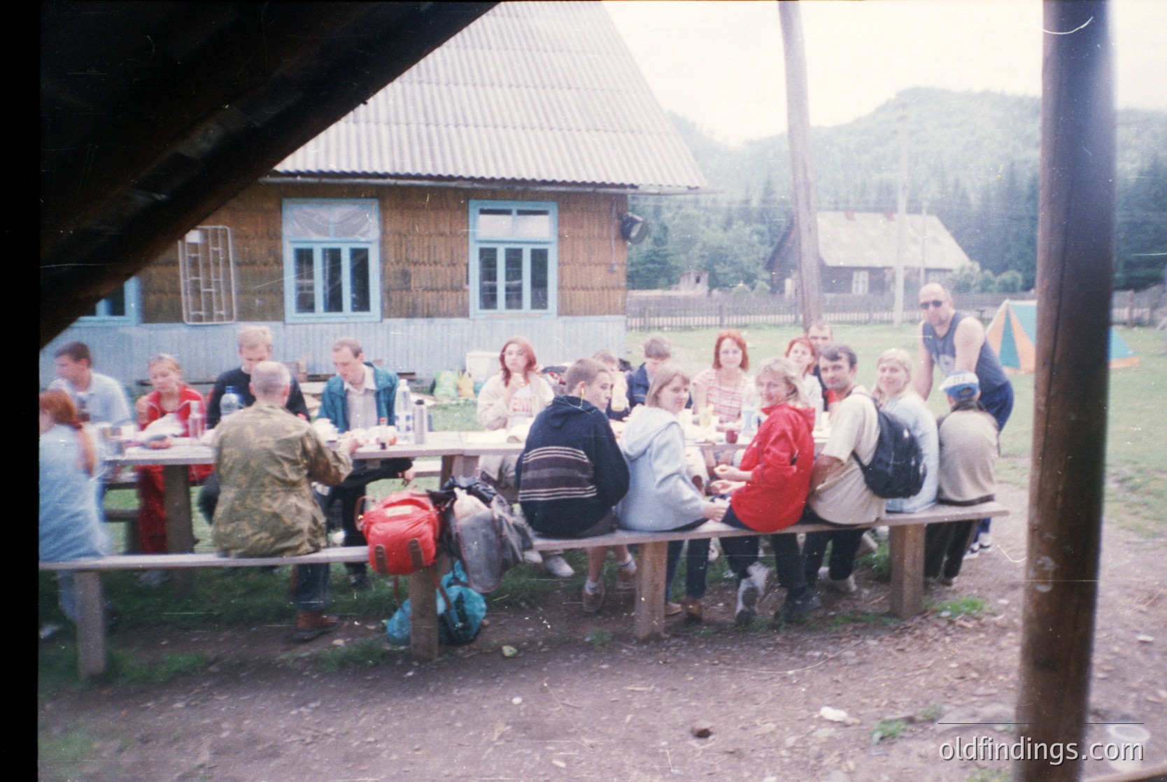 Group picnic in rustic alpine setting, 1990s. Wooden cabin with corrugated roof and metal-framed windows in background. Long picnic table surrounded by 15+ people—mixed genders, casual attire (backpacks, jackets). Mountainous terrain visible beyond.