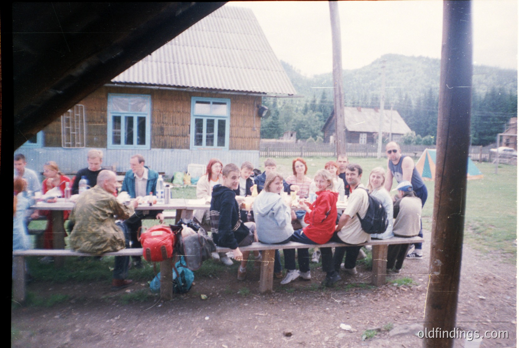 Group picnic under rustic wooden shelter in mountainous rural setting. Wooden tables, mismatched chairs, and casual attire suggest 1990s–2000s era. Backdrop features simple wooden houses with corrugated roofs and lush greenery.